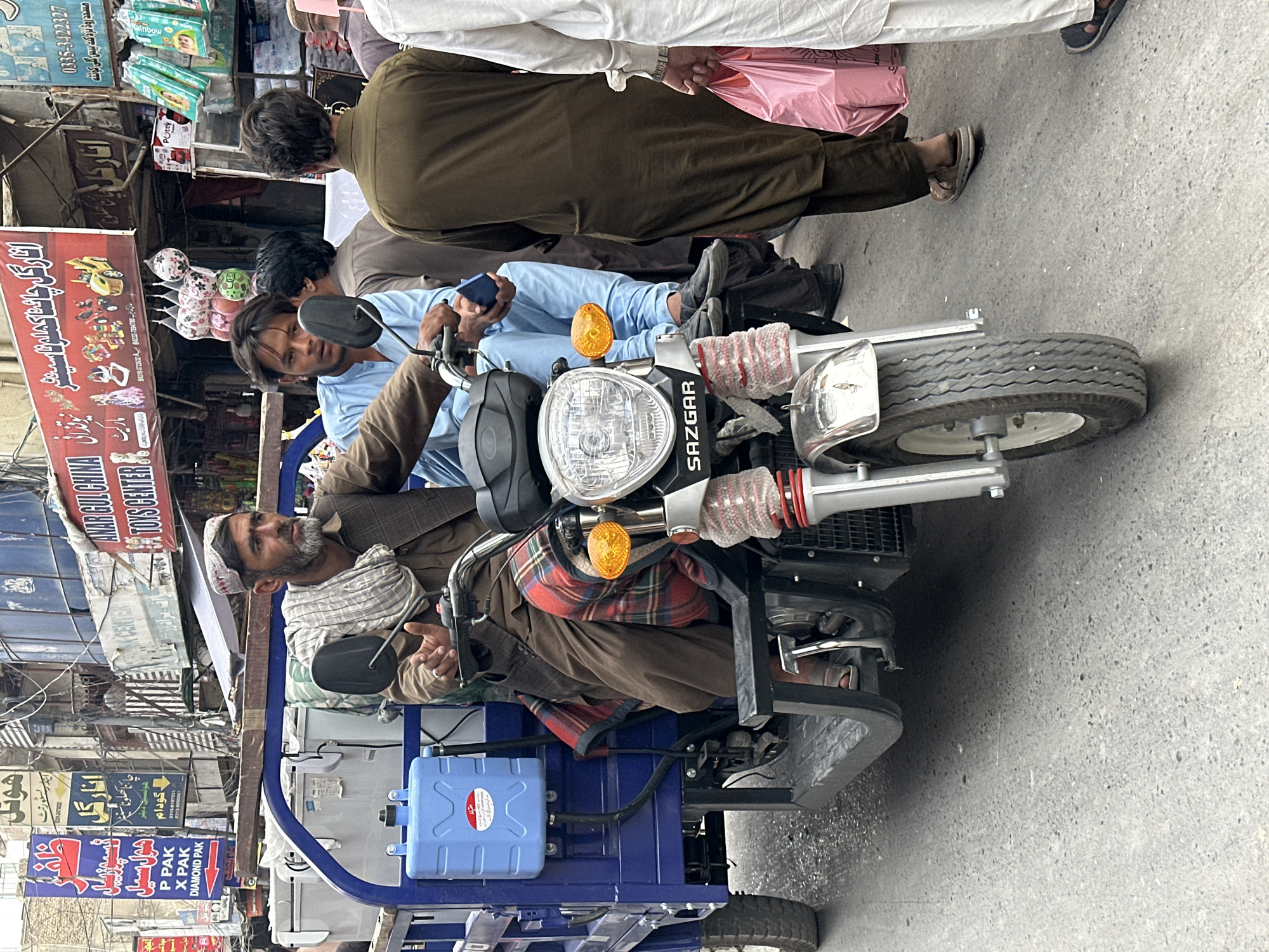This image depicts a street scene in Quetta City Tehsil, Pakistan. In the foreground is a Sazgar rickshaw with two men on it. The driver, an older man with a gray beard and a patterned head covering, is seated in the driver's position. A younger man, wearing a blue shirt, sits behind him, looking at a cell phone. Behind the rickshaw, there are people walking by on the sidewalk. Signs in Urdu advertise nearby businesses, including a toy store and a wholesale business. The scene appears to be taking place on a busy street.