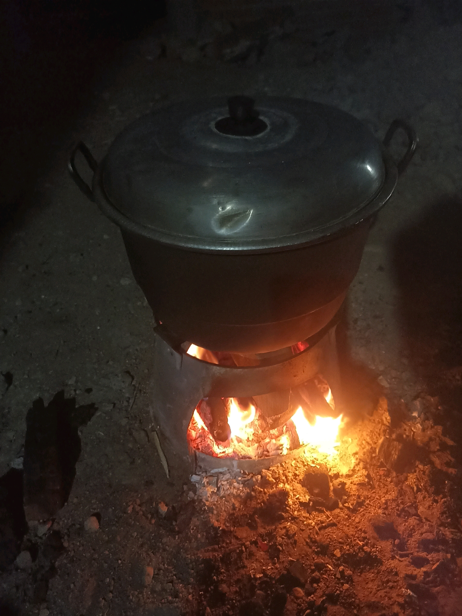 In Mojopanggung, Indonesia, a large, lidded metal pot sits atop a rustic, open-air wood-burning stove. The stove is made of metal with cutouts to allow for airflow and is actively fueled by a bright, flickering fire. Embers and ash are scattered around the base of the stove on the dirt and gravel ground. The pot appears to be cooking something, indicated by the heat radiating from the fire and the steam likely trapped beneath the lid. The overall scene depicts a traditional method of cooking, common in outdoor or less developed settings.