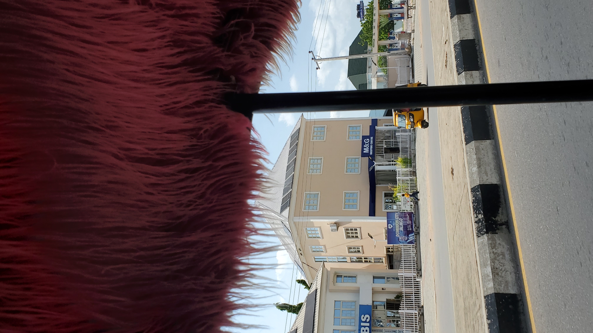 The image captures a street scene in Jalingo, Nigeria, viewed from inside a vehicle, possibly a car with a furry, red accessory obstructing part of the view. The perspective is tilted, suggesting the photo was taken from a moving vehicle.
The main subjects are buildings lining a paved street. One prominent building is a light-tan, multi-story structure with many windows, identified by signage as "M&G SHOPPING CENTRE". Another building adjacent to it has a blue sign with white lettering, though only "BIS" is clearly legible. A bright yellow auto-rickshaw is parked on the side of the road near the shopping center. A pedestrian, dressed in a yellow vest and dark pants, is walking on the sidewalk.
The scene depicts an ordinary day. The sky is blue with some clouds, indicating fair weather. The time of day appears to be daytime, likely mid-morning or afternoon, given the sunlight casting shadows. The presence of a shopping center, road infrastructure like curbs and a yellow line marking the edge of the road, and pedestrian activity suggest an urban or semi-urban environment.
Notable details include the textured, fuzzy red material in the foreground, which frames the view. The buildings showcase modern architecture with numerous windows and what appear to be solar panels on the roof of one building. Signage is a key detail, with "M&G SHOPPING CENTRE" and "DOMINION CITY JALINGO" visible. The overall impression is a snapshot of daily life and commerce in Jalingo.