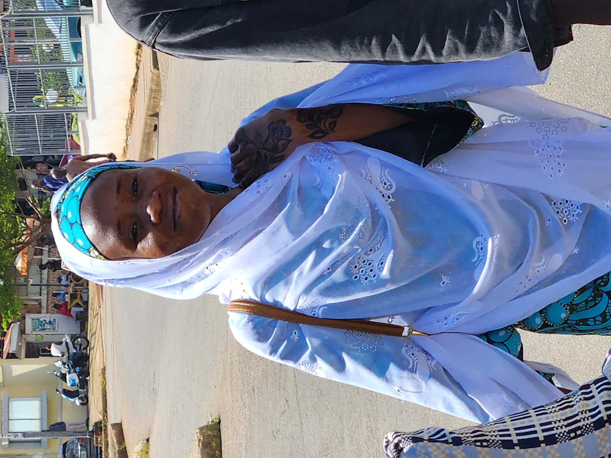 A medium shot captures a woman from the chest up, looking directly at the camera with a slight smile. She wears a white headscarf with a blue and patterned trim, and a white outer garment with intricate white embroidery. A brown purse strap is visible on her shoulder, and her left hand, adorned with henna tattoos, rests on her chest. To her right, a person in a dark, textured jacket is partially visible. The background shows a street scene with a paved road, a sidewalk, and a building with a light yellow facade. Several motorcycles are parked near the building, and in the distance, more people are walking and standing. The setting appears to be an urban environment under daylight.