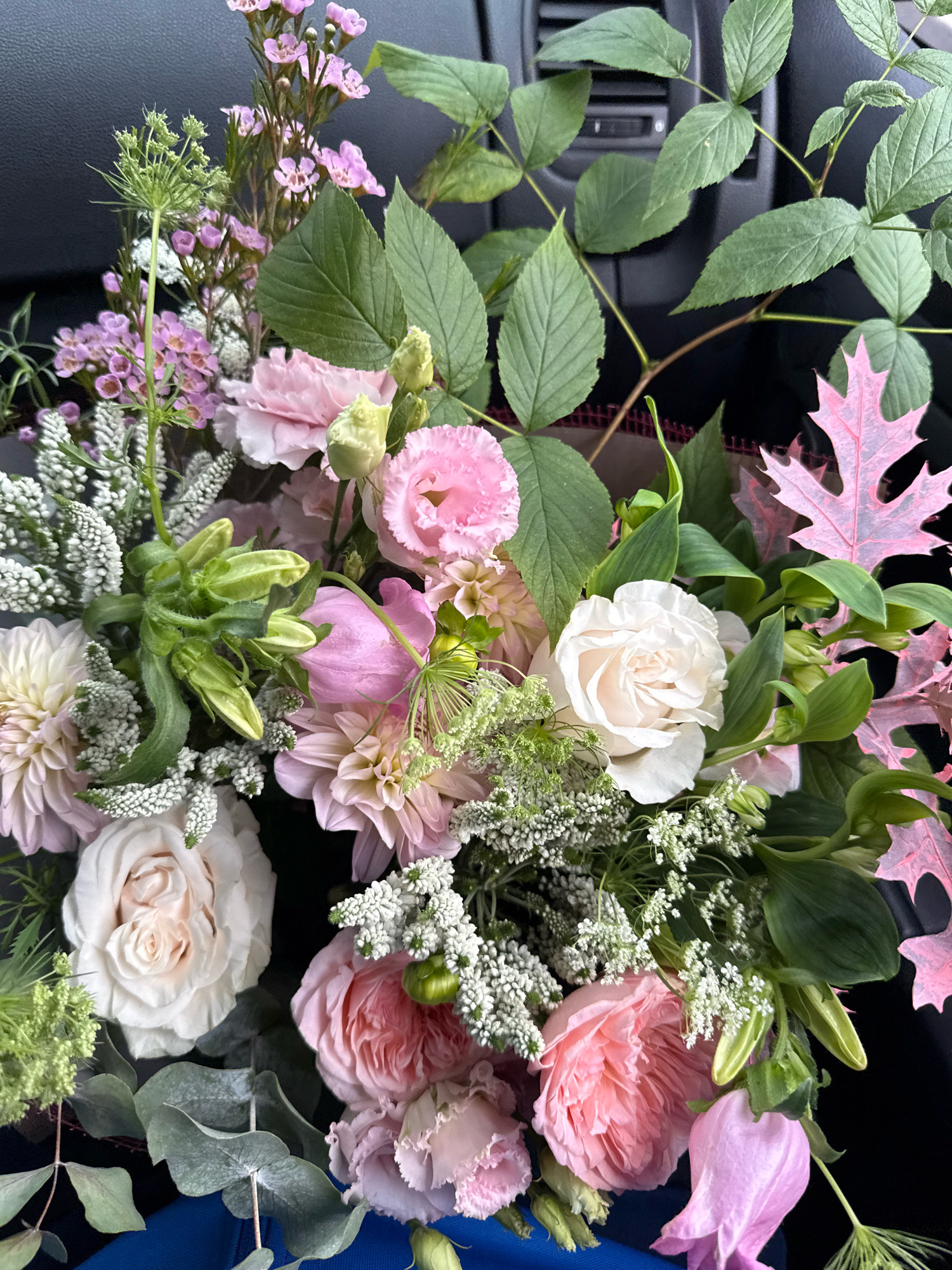 A bouquet of flowers, primarily in shades of pink and white, is arranged in what appears to be the interior of a vehicle. The background shows dark gray surfaces, suggestive of car interior components like a dashboard and air vents. Various floral elements are visible, including roses, carnations, dahlias, and smaller white filler flowers, interspersed with green foliage and branches. There are also distinctive pink leaves with prominent veining, possibly ornamental or decorative additions to the bouquet. The lighting suggests an indoor setting, likely daytime, given the natural light illuminating the flowers. No people are present in the image. The composition is a close-up view of the floral arrangement, filling the entire frame. There is no indication of the city of Tallinn, Estonia, or any specific urban environment.