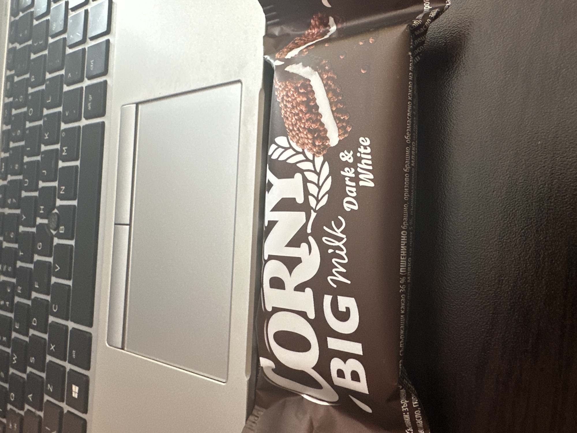 The image presents a close-up view of a laptop with a Corny Big Milk Dark & White chocolate bar resting beside the trackpad. The laptop keyboard is visible on the left side of the frame, showing a standard layout with keys such as "Z", "W", "X", "C", "V", "B", "N", "M", among others. To the right, the chocolate bar is prominently displayed with its packaging partially visible, clearly showing the brand name "Corny Big Milk" and the flavor "Dark & White". The chocolate bar seems to be sitting on a dark, smooth surface, likely a table. There are some words printed in Bulgarian on the packaging, including the phrase "ПШЕНИЧНО брашно," which translates to "wheat flour". The location in Sofia, Bulgaria can be inferred based on the Bulgarian text on the chocolate packaging.