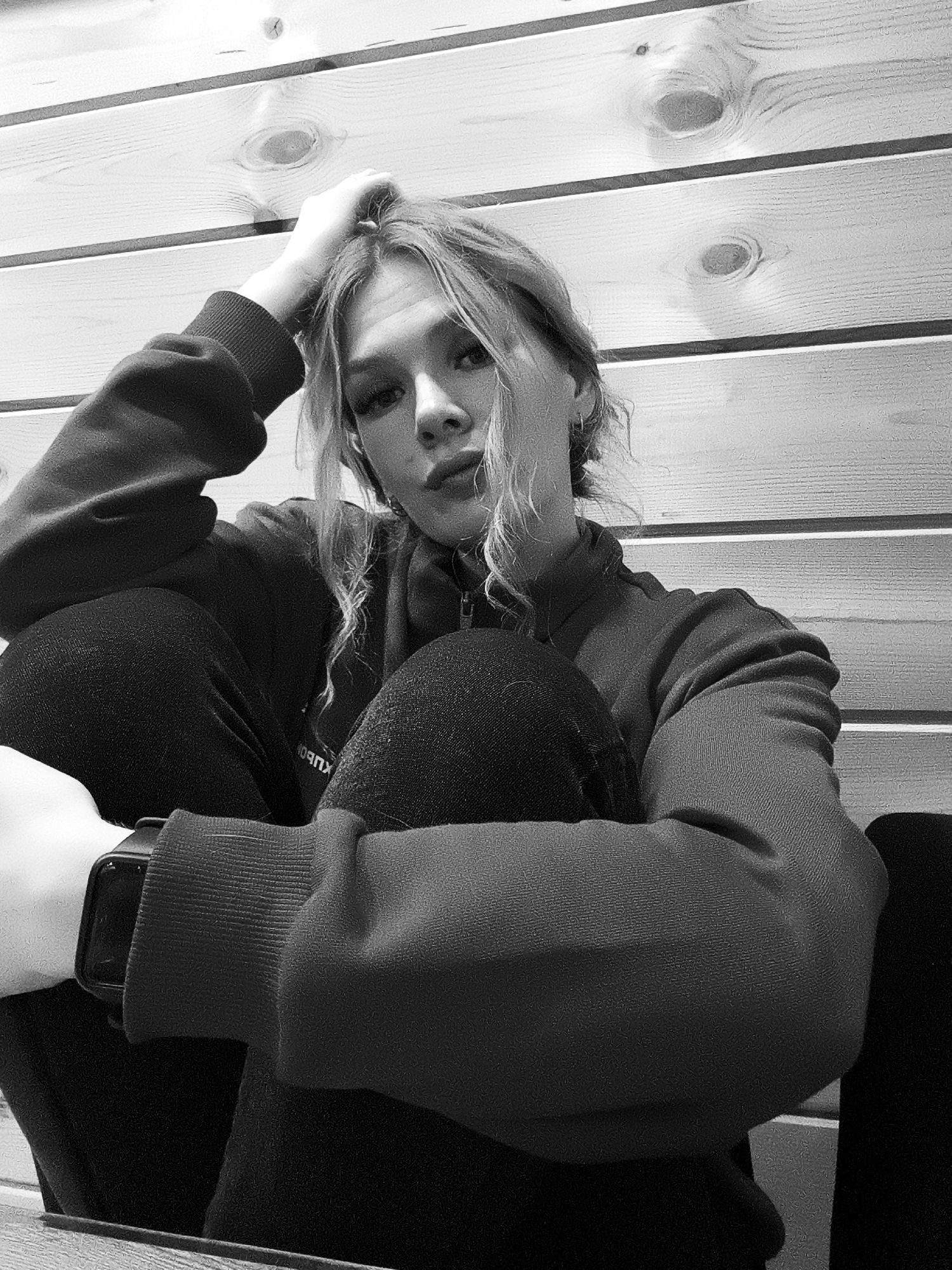 A black and white indoor photograph depicts a young woman sitting cross-legged, her knees pulled up to her chest. She is wearing a long-sleeved zip-up shirt or jacket and dark pants. Her left hand is resting on her head, and her right arm is wrapped around her legs. A smartwatch is visible on her left wrist. The background is a wall constructed of horizontal wooden planks.
The image was taken in Русский Вожой, Russia.