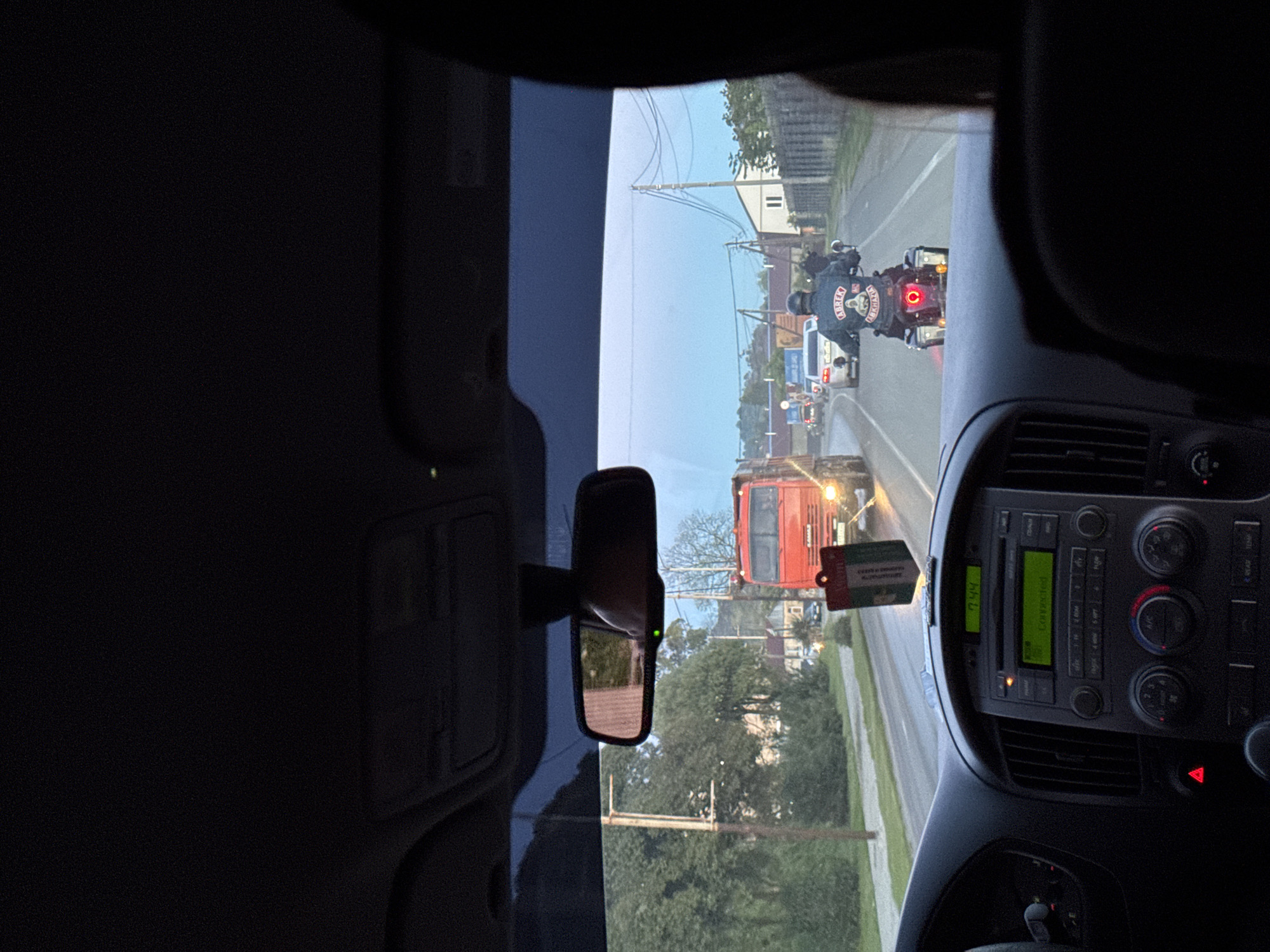 From inside a vehicle, a street scene is viewed. A truck is directly ahead, followed by other vehicles. A motorcycle with two people is positioned behind the other vehicles. The rider wears a jacket with distinctive emblems. Buildings, power lines, and trees line the street. The vehicle's dashboard and rearview mirror are in view. The location appears to be the city of Tkhubyn in Abkhazia, inferred from vehicles and landscape.