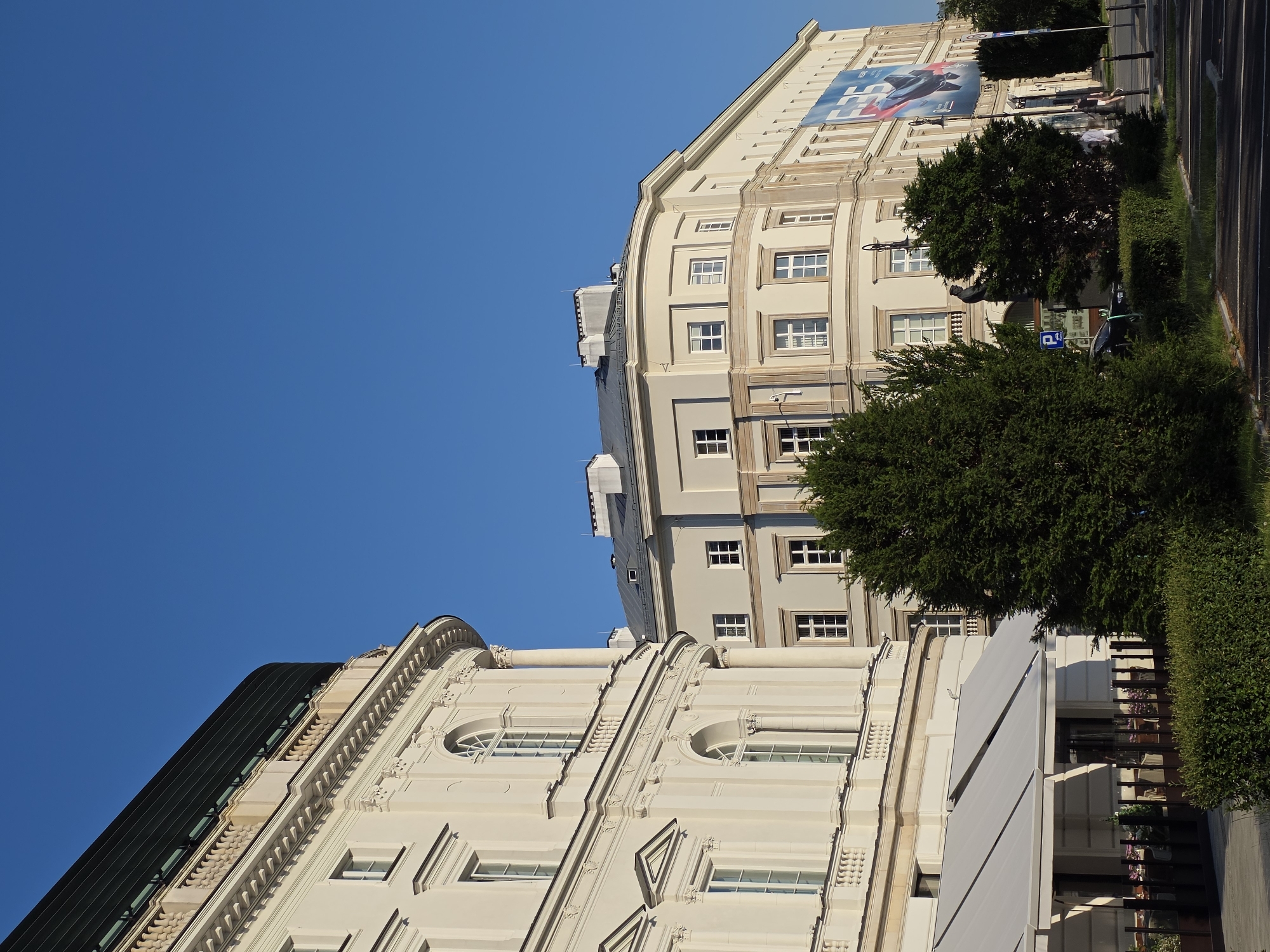 This is a photograph of the Warsaw University of Technology in Warsaw, Poland. The image shows the main building of the university, which is a large, ornate building with a light-colored facade. The building has many windows, some of which are arched. There is a banner on the side of the building that says "Warsaw University of Technology". The sky is clear and blue, and the sun is shining. In the foreground, there are some trees and bushes.