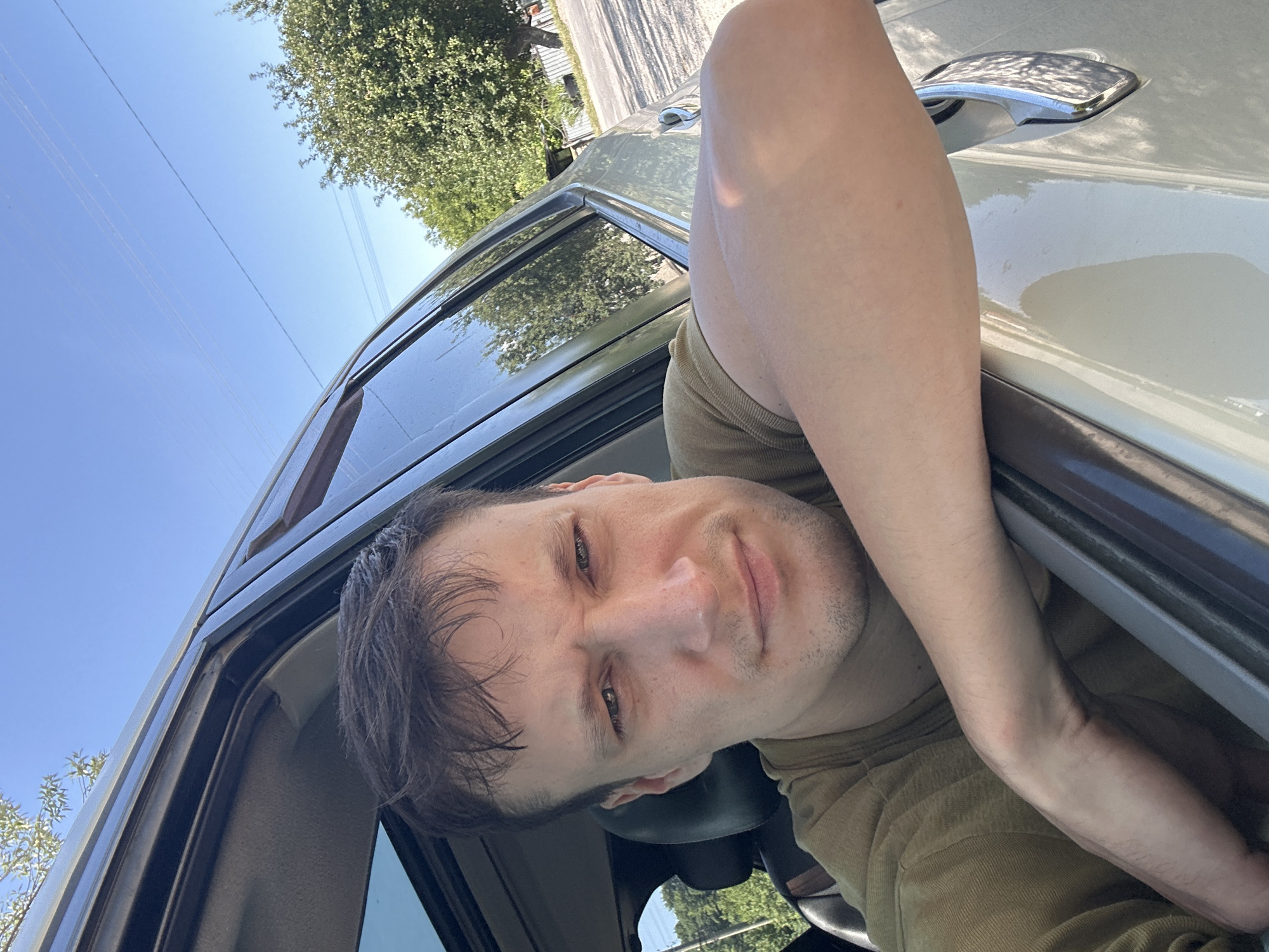 A man leans out of a car window, with his arm resting on the sill. He has short, dark hair and is wearing a green t-shirt. He is looking directly at the camera. The car appears to be parked on a paved road, with trees and a fence visible in the background. The sky is clear and blue. The scene is likely located in Makiv, Ukraine.