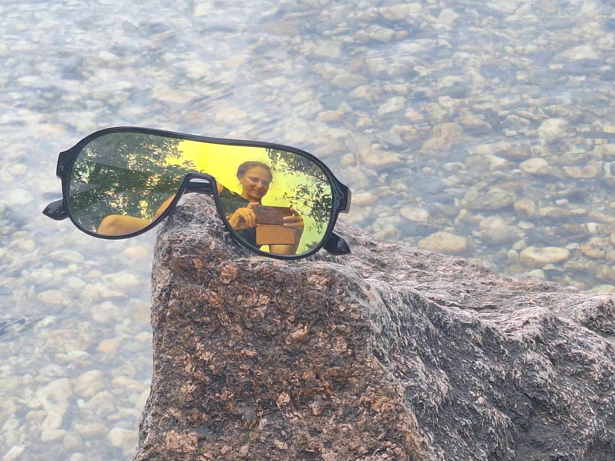 A pair of large, black-framed sunglasses with yellow lenses rests on a rough, grey and brown granite rock. The sunglasses are angled to reflect a scene of a person holding a phone, taking a selfie, and trees with green leaves. The rock is situated on the edge of a body of clear water filled with small, smooth, beige and grey stones. The background shows more of the stony waterbed under the surface. No specific Leipzig landmarks are identifiable in this image.