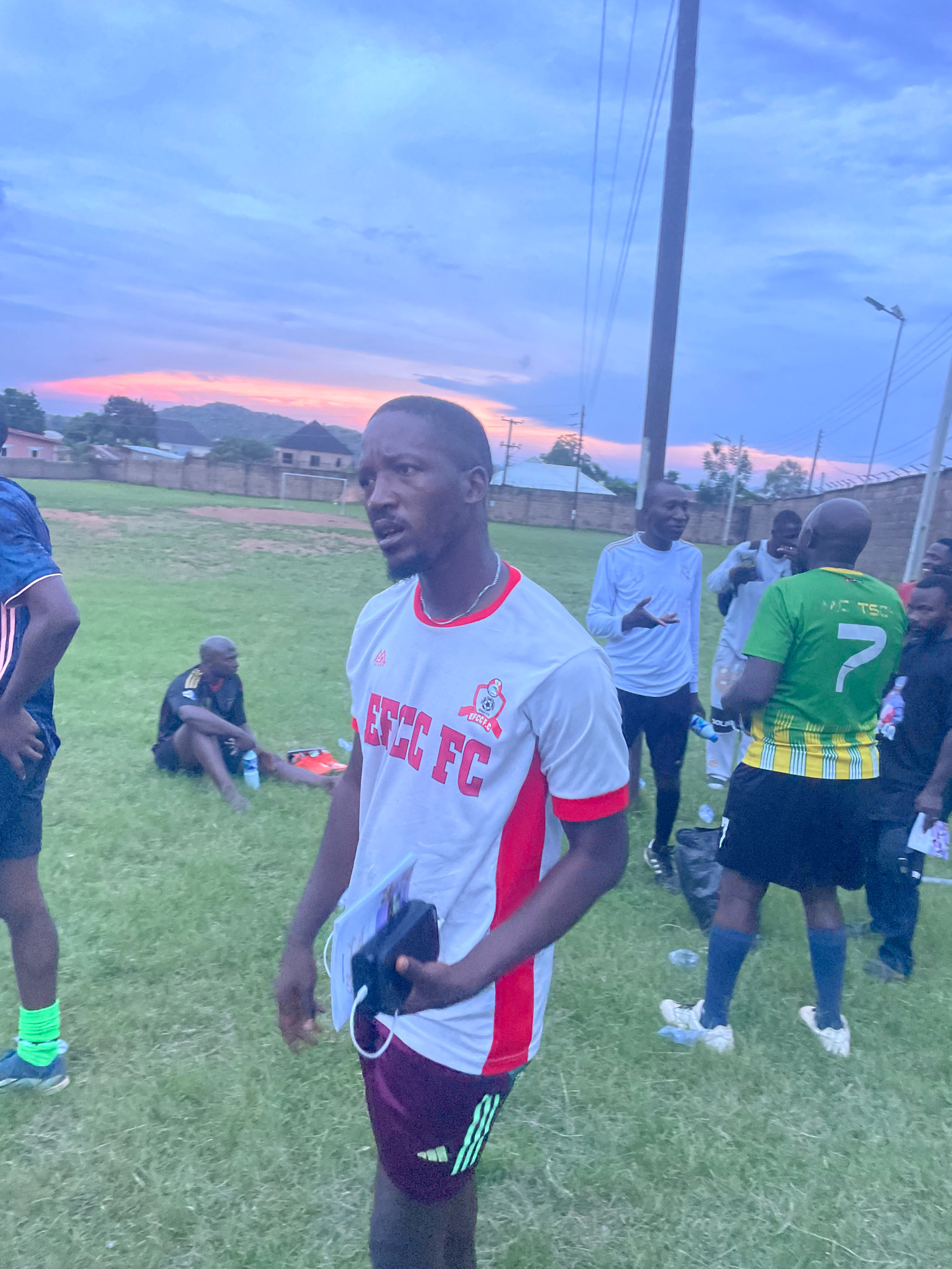 The image depicts an outdoor scene at dusk, likely in Jos, Nigeria. A group of men, possibly a soccer team, are gathered on a grassy field. The sky exhibits a gradient of colors, from a pale blue at the top to hues of pink and orange near the horizon, suggesting sunset. In the foreground, a man wearing a white and red soccer jersey with "EFCC FC" printed on it is looking to his right and holding a black electronic device and a folder. Behind him, other men are also dressed in athletic wear. One man in a green jersey with the number 7 on it is visible, and another is seated on the grass. In the background, there are buildings and hills, characteristic of the Jos Plateau. The overall atmosphere appears to be casual, possibly after a practice or game.