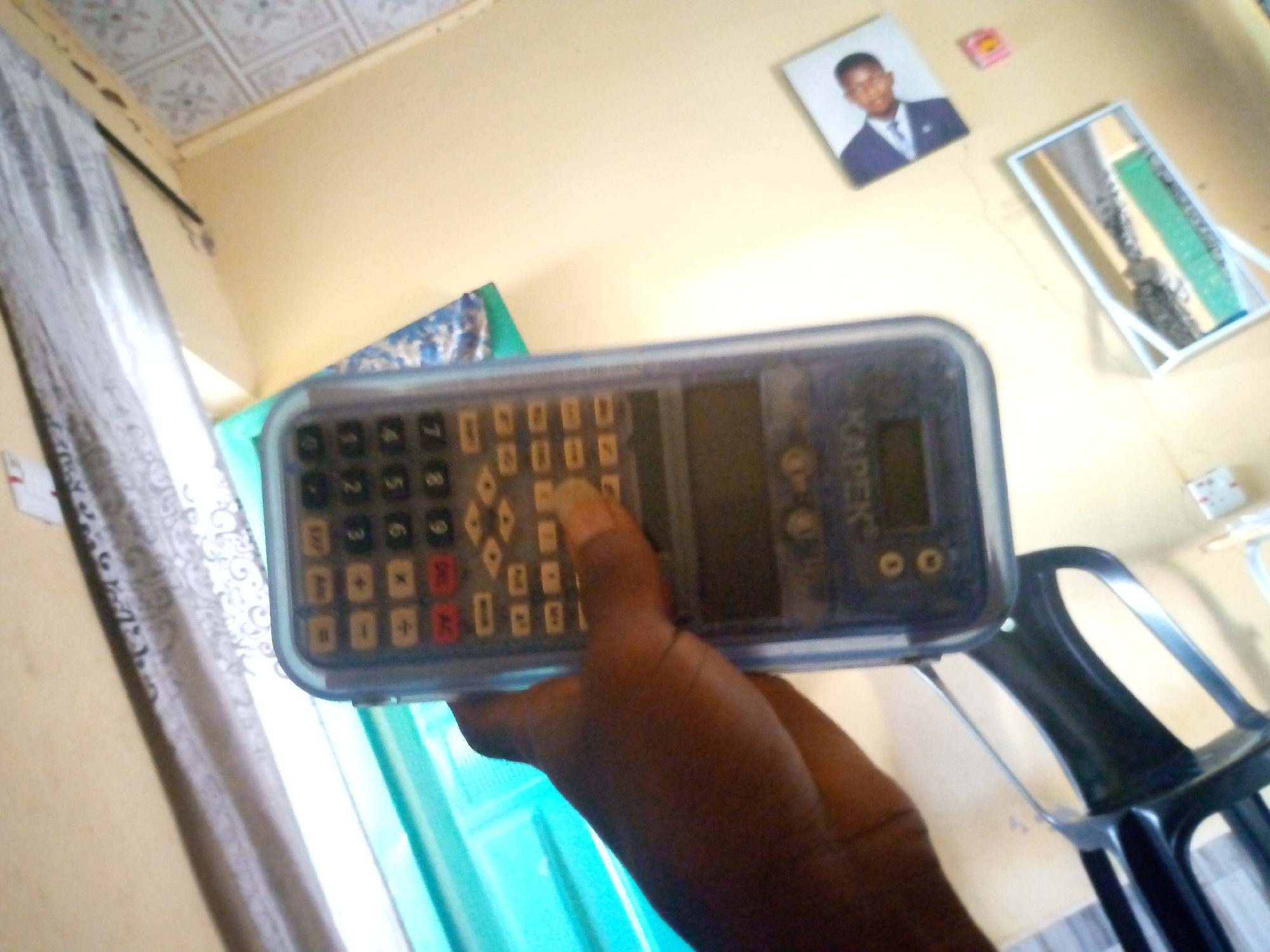 The image depicts an indoor scene, likely a room in Jalingo, Nigeria. In the foreground, a hand holds a dark-colored calculator with a clear plastic cover. The calculator has a numeric keypad with buttons for digits 0-9, as well as operation symbols and other function keys. The display screen of the calculator is visible, showing some faint markings. The background of the image reveals a wall painted a light yellow color. On the wall, there are a few framed items: a small framed photograph of a young Black man in a suit and tie, and a rectangular mirror with a white frame, reflecting some of the room's interior. A partially visible curtain with a grey and white pattern hangs to the left of the frame. To the right, a black plastic chair with a curved backrest is visible. The ceiling appears to be paneled with a patterned design. The overall lighting suggests it's daytime, possibly with natural light entering from a window, indicated by the bright area on the left and the cyan-colored vertical blinds.