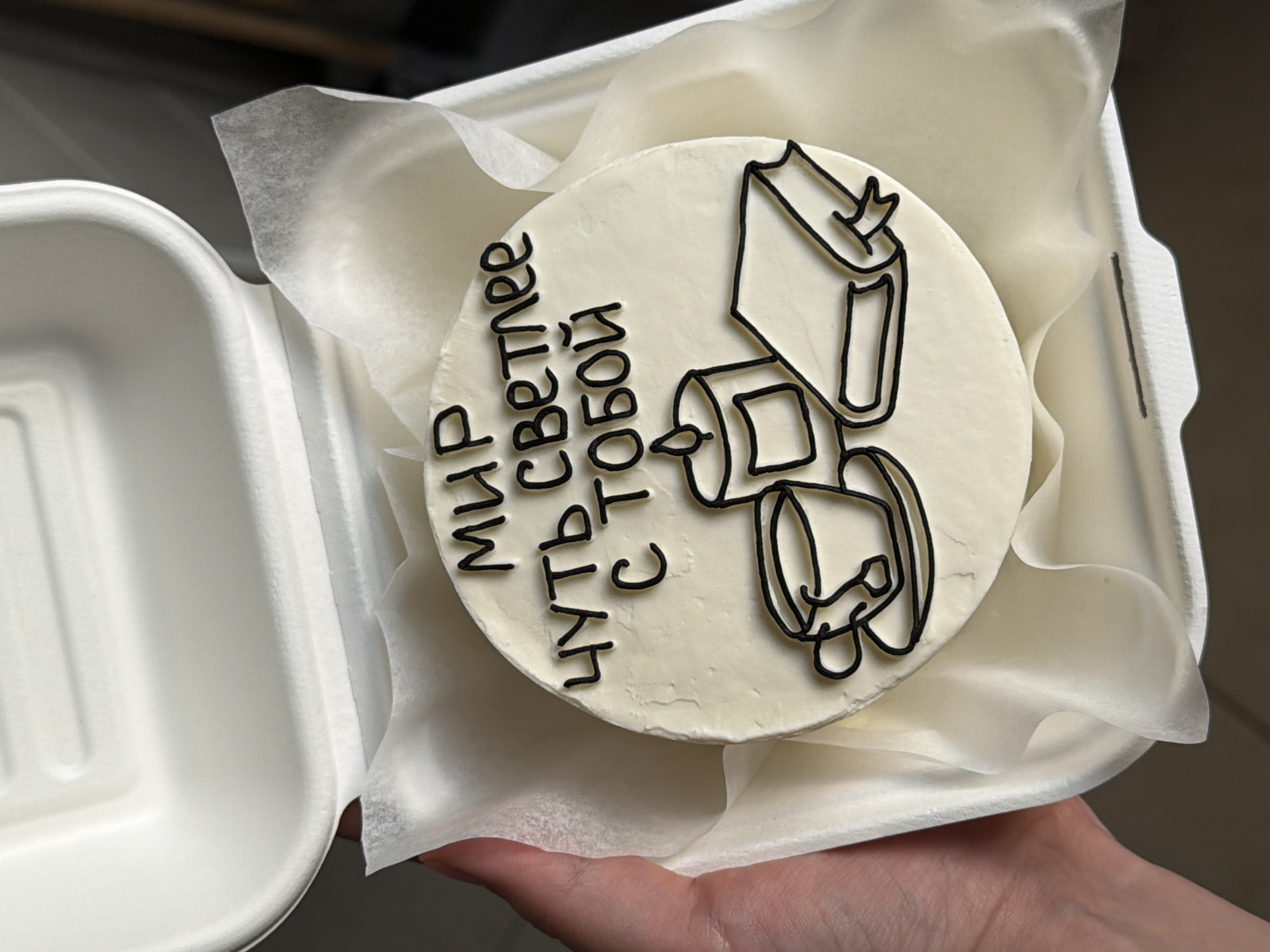 A top-down shot depicts a small round cake decorated with black-outlined drawings and lettering, nestled inside a white, biodegradable food container. The cake sits on a bed of crinkled white tissue paper that cushions it within the container. The black drawings on the cake seem to depict stacked objects, possibly books or abstract shapes. To the left of the objects, vertical lettering in black is discernible, although the language is not immediately identifiable as English.
The food container appears to be made of a molded fiber material, similar to sugarcane pulp. Part of the container's lid is visible on the left side of the frame. A human hand with pale skin is holding the container, with the fingers of the hand subtly visible along the bottom right edge of the image.
The backdrop appears to be simple and uncluttered, suggesting an indoor setting.
The location details of Tel-Aviv, Israel were not determinable in this image.