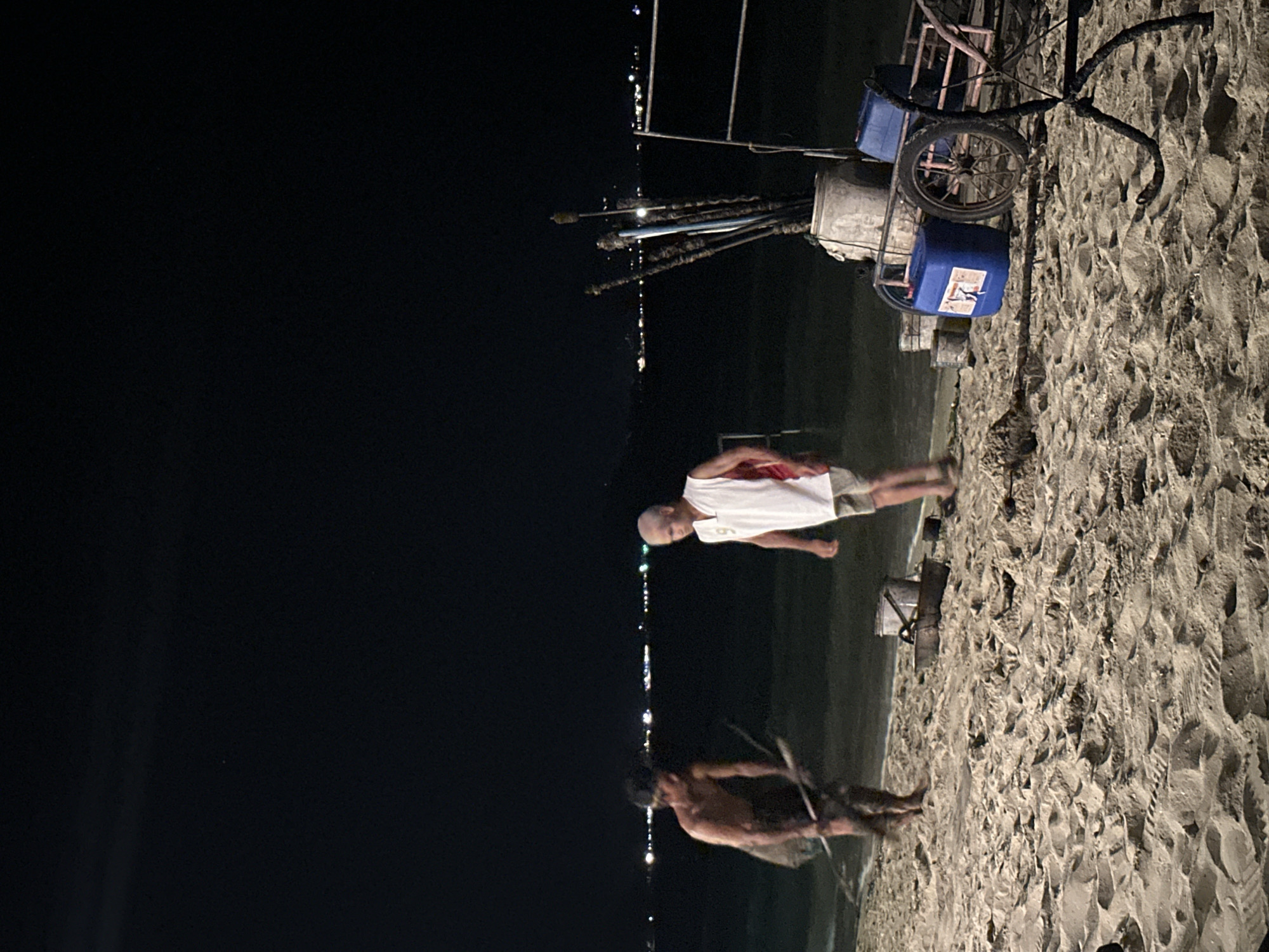 At Baan Klong Prao, Thailand, the scene features two people on a sandy beach at night. One man, wearing a white shirt and shorts, stands upright. The other, shirtless and facing away, appears to be working with long sticks. Behind them is a cart carrying supplies, including blue containers and what appears to be fishing equipment. The background is a dark sky with a few distant lights that appear to be city lights. The beach has distinct footprints and patterns in the sand.