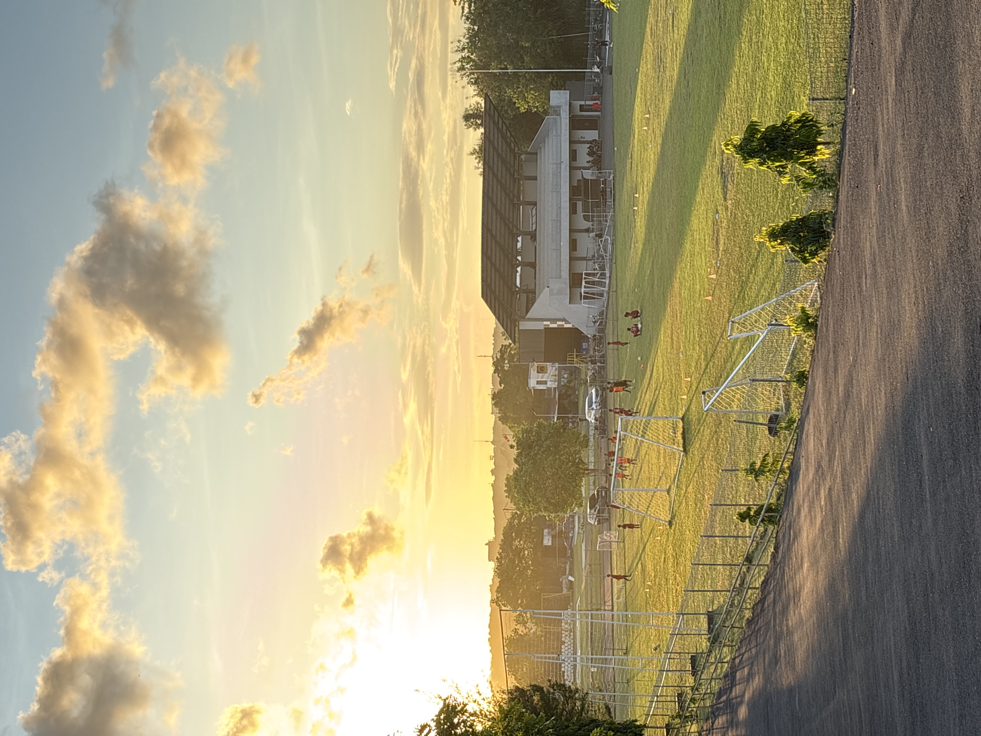 The image depicts a soccer field during a sunset in Kuta Selatan, Indonesia. The sky is filled with clouds illuminated by the setting sun. A multi-story building with a partial roof structure is visible in the background. Several individuals, possibly soccer players, are distributed across the field. Soccer goals are in place. The field is bordered by a fence and a paved path. The overall scene is bathed in warm, golden light.