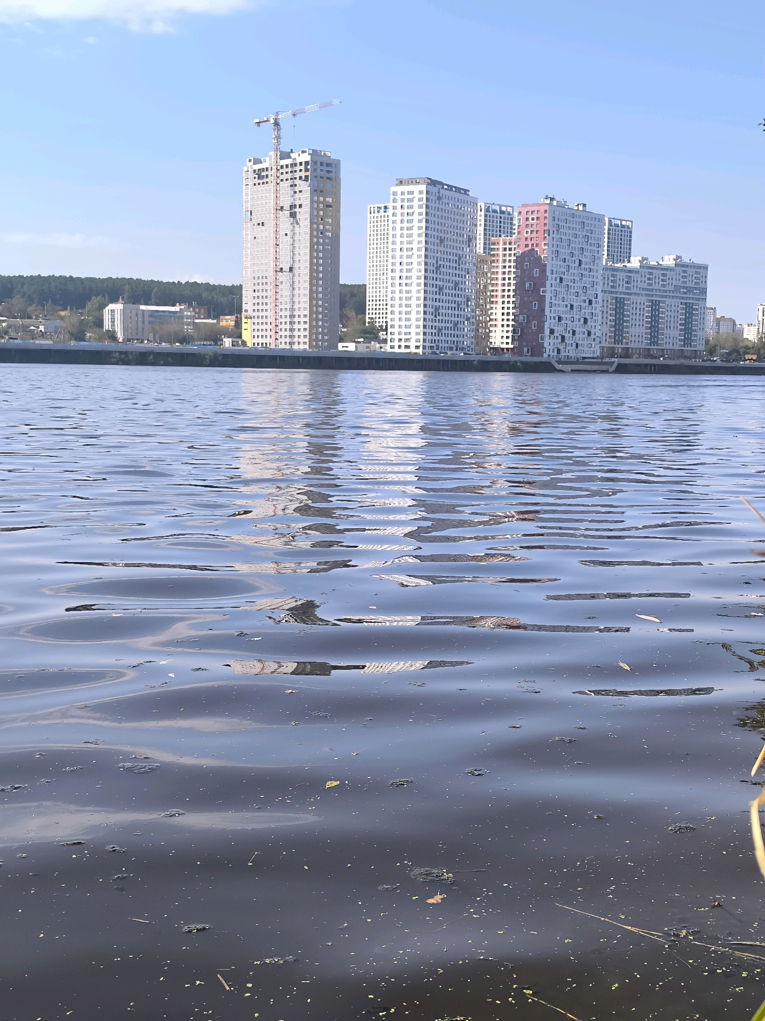 This image shows a body of water in the foreground with a city skyline in the background. The skyline features several tall, modern buildings, some of which are under construction. A prominent construction crane is visible next to one of the taller buildings. The water in the foreground is choppy, with reflections of the buildings visible on its surface. There is also debris floating on the water. The sky is blue with some clouds. The city of Yekaterinburg, Russia is known for its riverside development, and this scene is likely of the Iset River, which flows through the city. The modern architecture and ongoing construction suggest an urban area undergoing development.