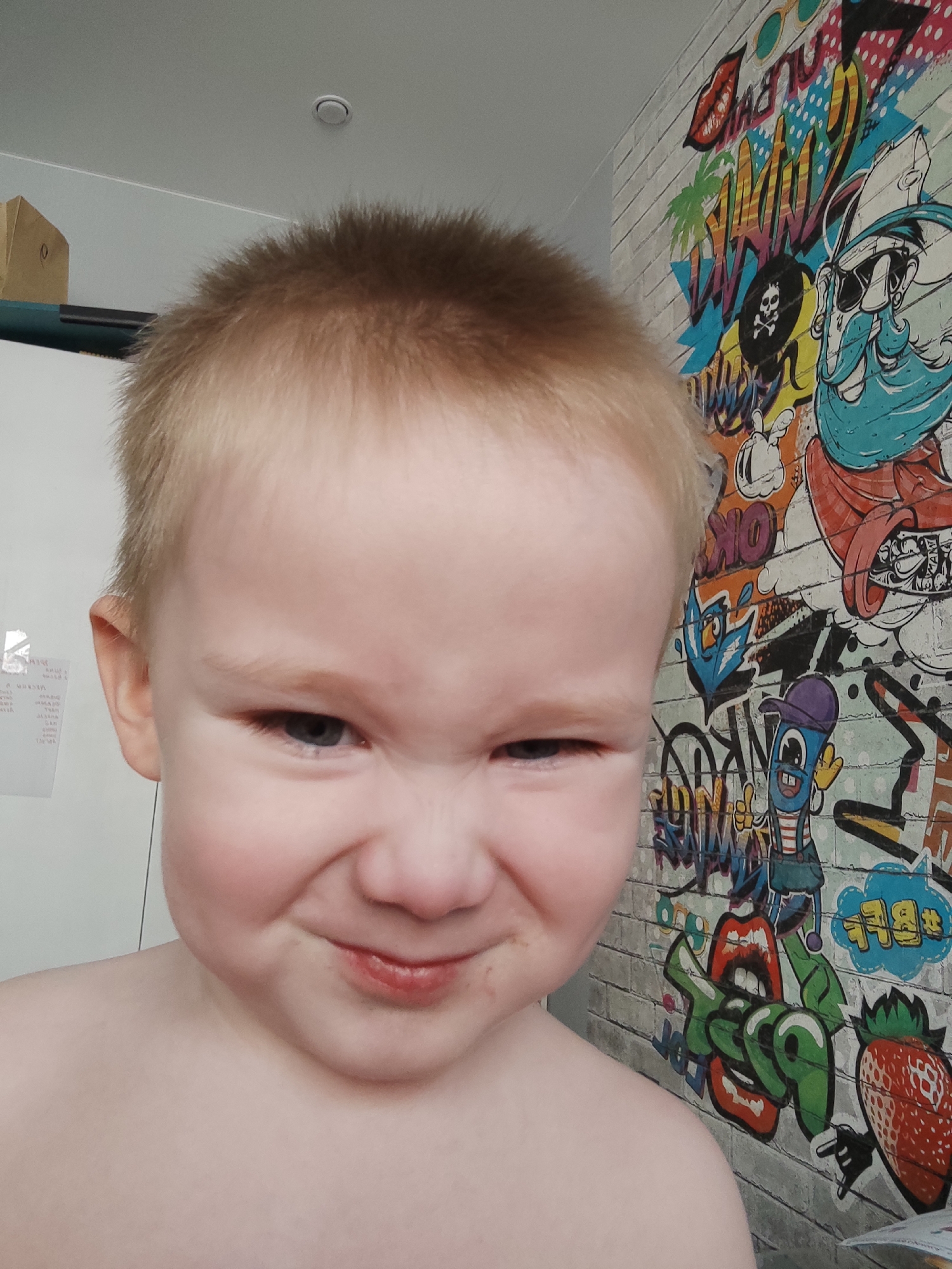 The image captures a close-up portrait of a young, fair-skinned child with short blonde hair and blue eyes, likely taken indoors in Izhevsk, Russia. The child looks directly at the camera with a playful, mischievous expression, eyes squinted and a slight, lopsided smile. Their bare shoulders and chest are visible, suggesting an informal or relaxed setting.
The background is divided into two distinct sections. To the left, a plain white wall features some white papers with unreadable text and a cardboard box with the number "0" visible on its side. To the right, a vibrant and busy wallpaper covers what appears to be a brick-patterned wall. This wallpaper is adorned with colorful graffiti-style art, depicting various cartoonish and pop-art elements. Notable features in the graffiti include a blue-bearded character wearing sunglasses and a pirate hat, a skull and crossbones, stylized red lips, a palm tree, a red strawberry, a blue bird, and abstract shapes and lettering. Visible words on the wallpaper include "DAMN" and "OK." Above, a white ceiling with a single, recessed circular light fixture is partially visible. The lighting is bright and even, indicative of daytime.