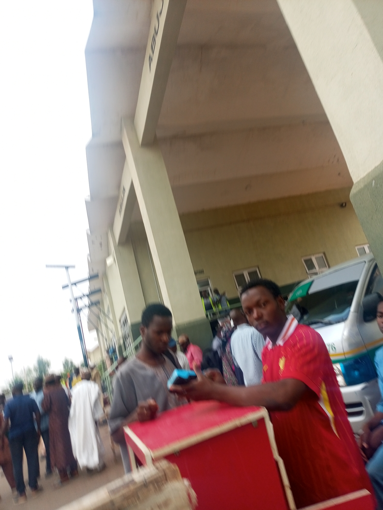 The image depicts an outdoor scene at Abuja, Nigeria. There are several individuals present, dressed in various attires. In the foreground, one man wearing a red shirt leans on a red structure with a white trim. Beside him, another man holds a small electronic device. In the background, a diverse group of people can be seen standing and walking near a building with the word "ABUJA" visible on a large structural beam. An ambulance is also present.