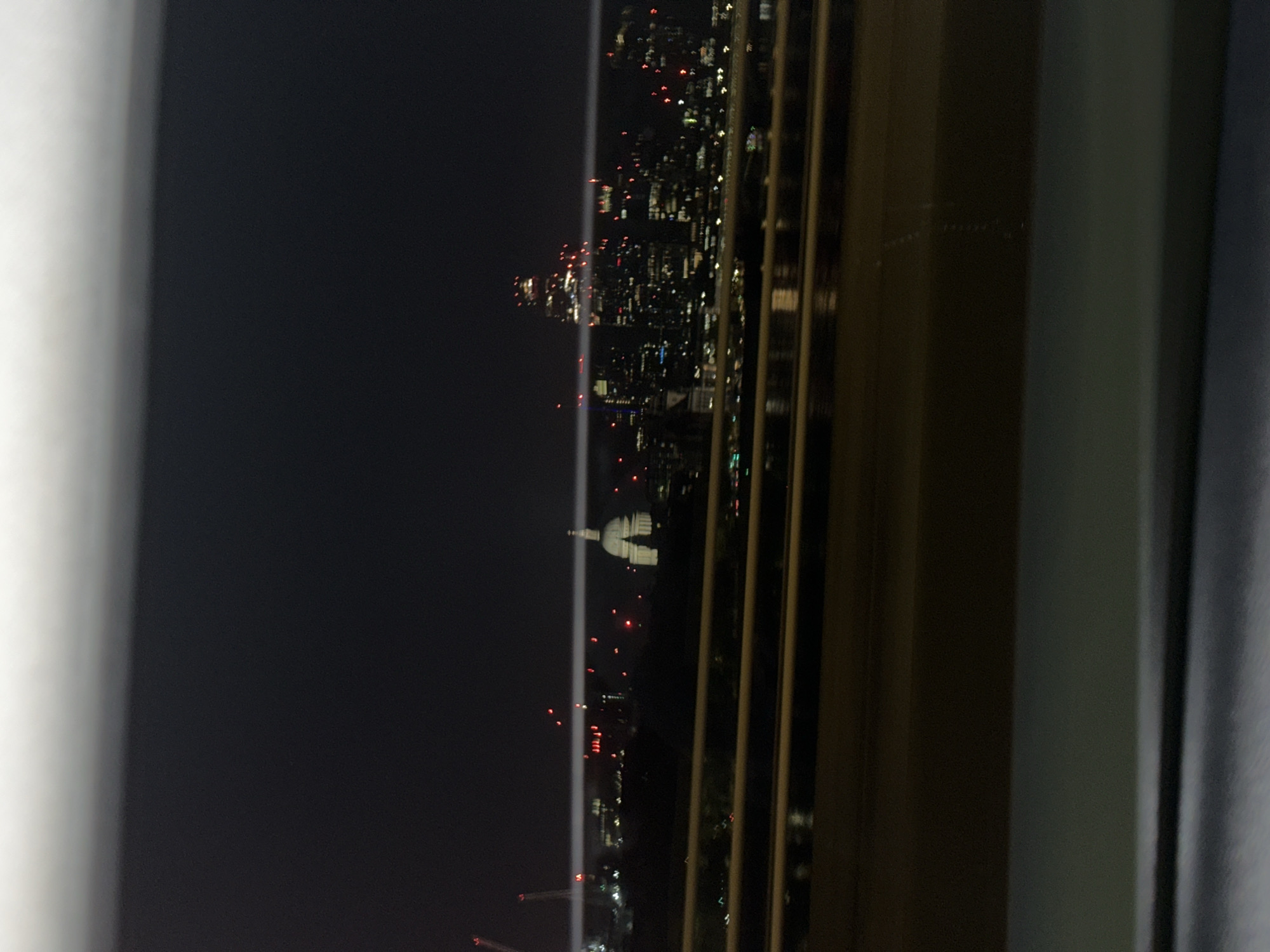The image depicts a nighttime view of London, United Kingdom, from a high vantage point, possibly through a window. Prominently visible is St. Paul's Cathedral, identifiable by its iconic dome, illuminated against the dark sky. The city lights stretch into the distance, interspersed with the red lights of tall structures, potentially construction cranes or tall buildings. The foreground includes elements of the window frame or structure from which the photo was taken.