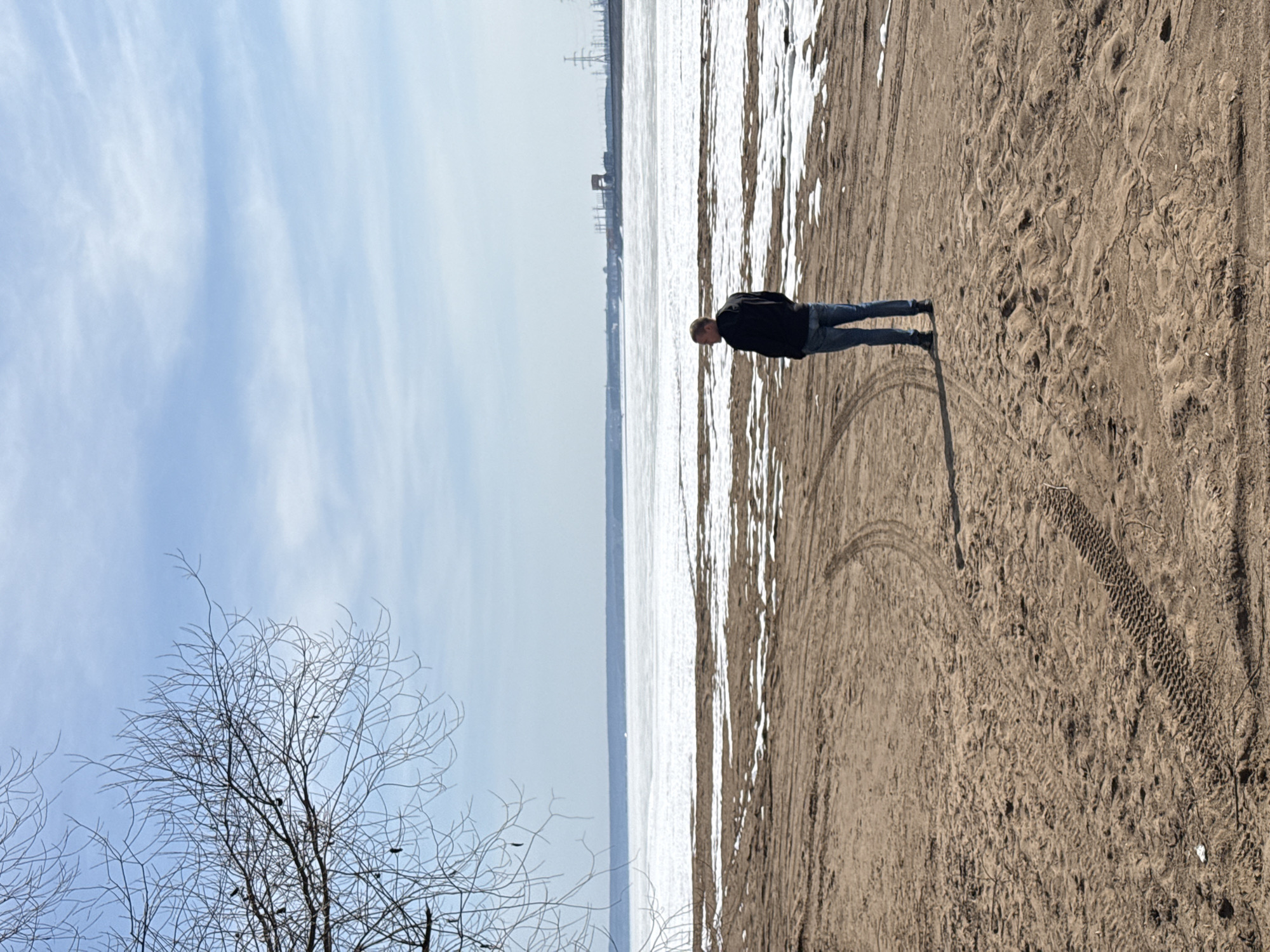 The image depicts a man standing on a sandy beach. The beach is marked with tire tracks and patches of snow. In the background is a frozen body of water, possibly a river or lake, with industrial structures visible on the opposite shore. A bare tree is in the lower left corner of the frame. The sky is partly cloudy. The location is likely near the city of Noviy, Russia, based on the latitude and longitude, which have not been provided.