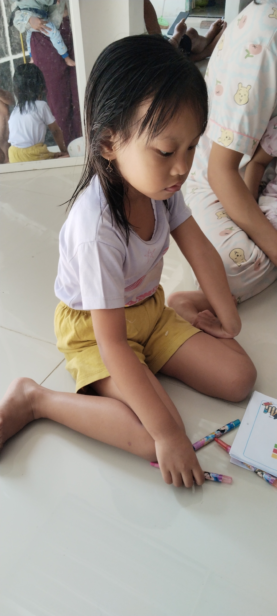 A young girl is sitting on a white tiled floor, wearing a light purple t-shirt and yellow shorts. She has dark, wet hair and is wearing a small gold earring. She is looking down and appears to be drawing or coloring with a set of colored pencils. Next to her is an open book or sketchbook with colorful illustrations. In the background, another person wearing pajamas with a Winnie the Pooh print is visible, and a reflection in a mirror shows another child being held. The scene suggests an indoor activity, possibly at home, in Kecamatan Pati, Indonesia.