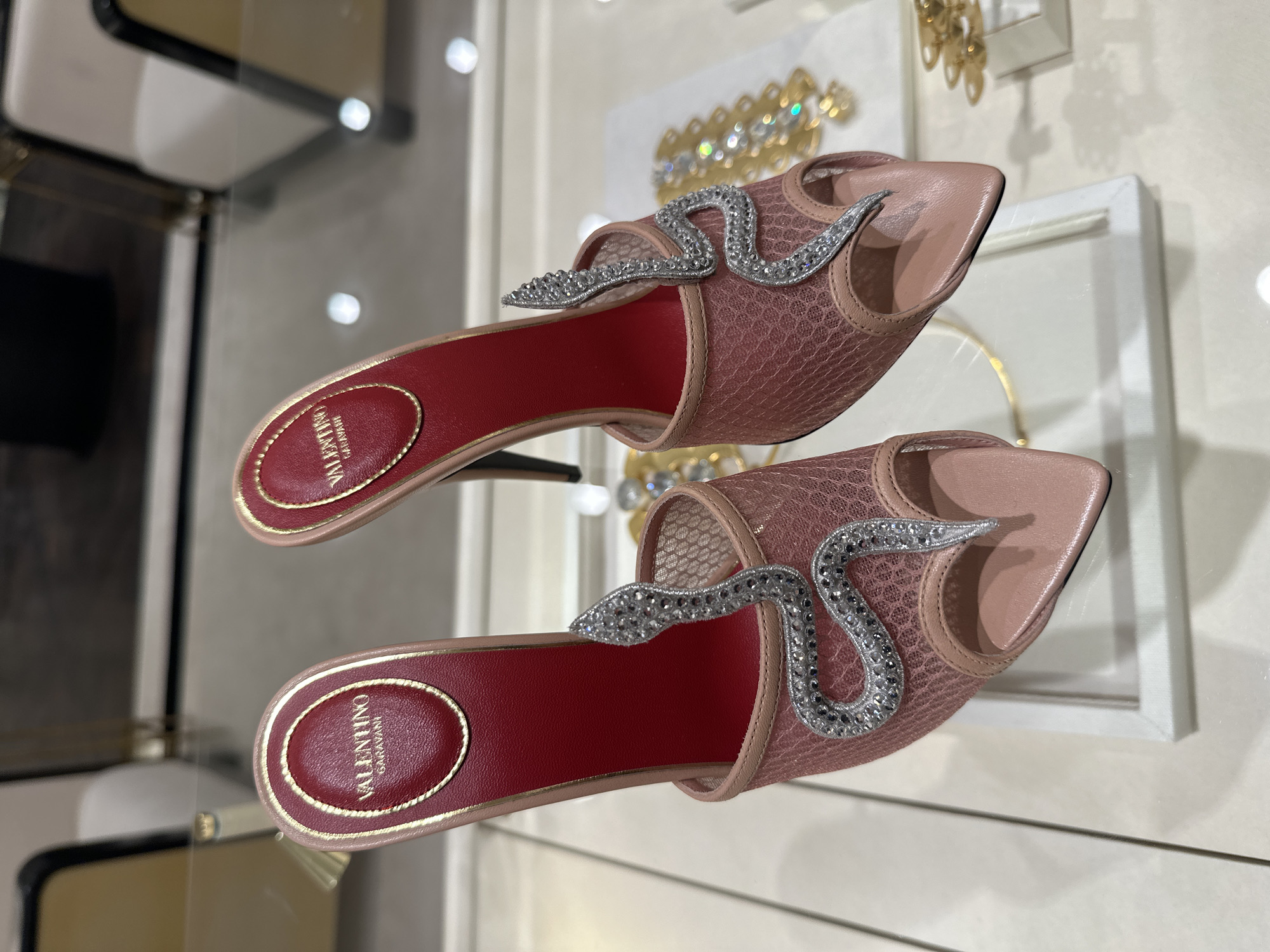 The image shows a pair of Valentino Garavani sandals displayed within a glass-fronted cabinet, likely in a high-end retail environment. The sandals are pink and feature a red insole with the brand name "VALENTINO GARAVANI" printed in gold. Embellishing the shoe's upper is a decorative snake motif adorned with small, sparkling stones. The sandals sit on a glossy surface, which reflects the shoes and surrounding elements, indicating a polished or mirrored display. The backdrop includes other items, possibly jewelry, also showcased within the cabinet. Although the exact location within London cannot be determined from the image, the presence of luxury items suggests a location in an upscale shopping district.