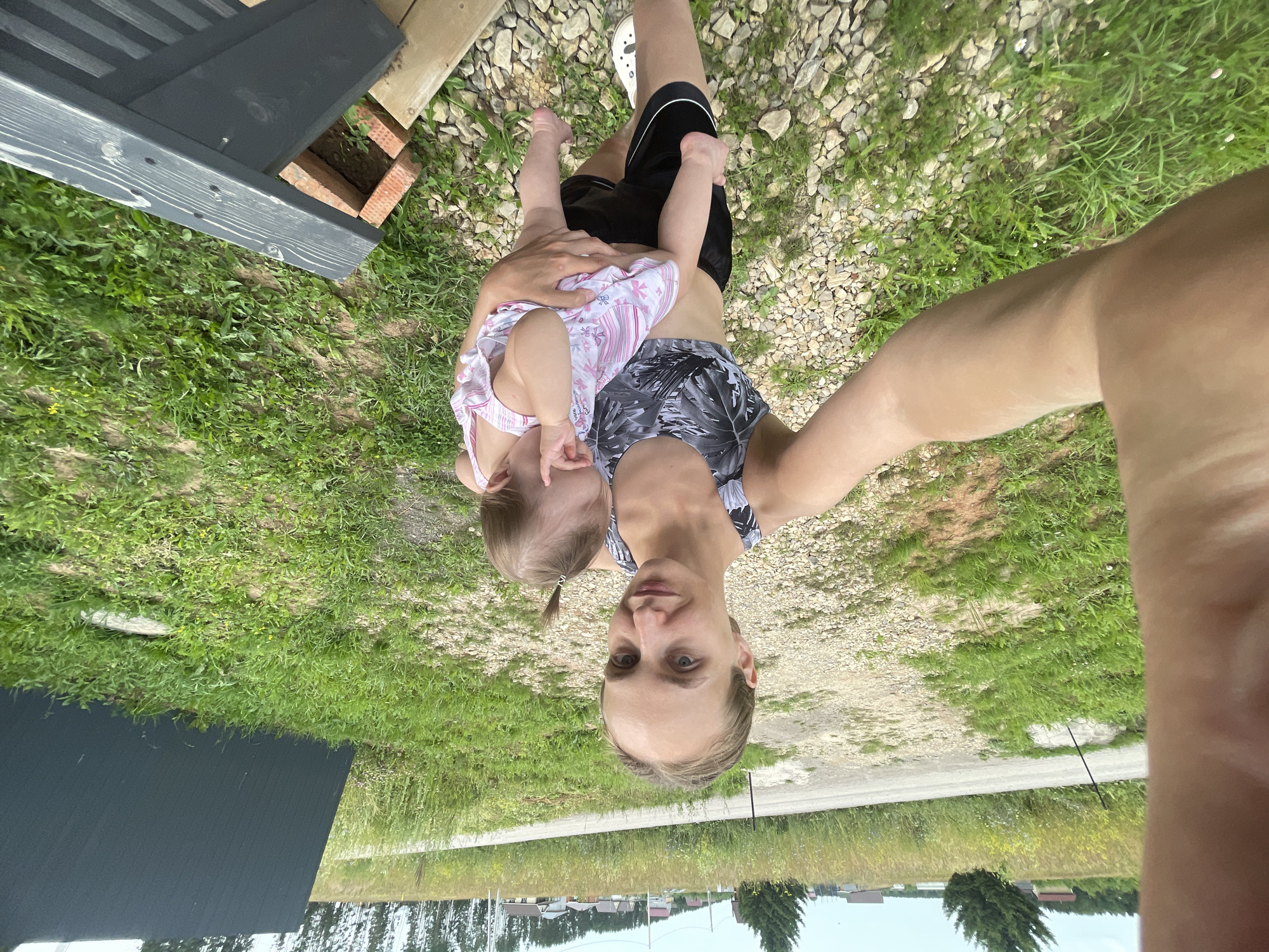 The image is a selfie taken outdoors, with the subjects oriented upside down from a standard perspective. A person is holding a baby in their arms. The person is wearing a patterned tank top and dark shorts with white trim. The baby is wearing a pink and white striped dress with a floral pattern. They appear to be in a natural setting, with grass, gravel, and some wooden structures visible. In the background, there is a glimpse of a rural landscape with houses and trees, suggesting a location in Russia, specifically Подшивалово. The lighting suggests it is daytime, likely sunny. The overall mood seems relaxed and casual, with the person taking the selfie. No visible text is present in the image.