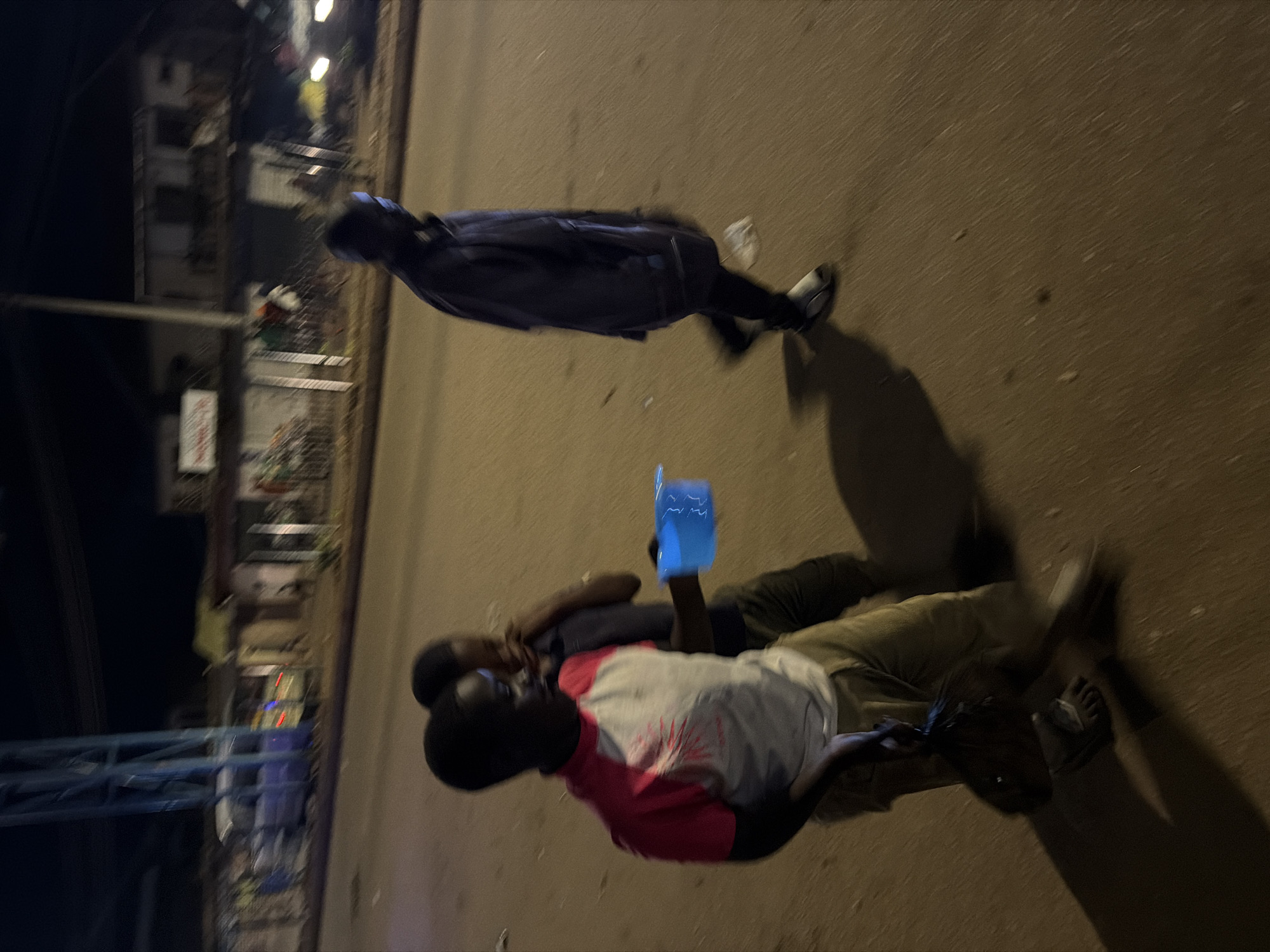 In Bukuru, Nigeria, at night, three individuals are walking on a paved road. In the foreground, a young person wearing a pink and white t-shirt and khaki pants holds a bright blue plastic container. To the left of this individual, another young person is partially visible, with their arm around the first person's shoulder. Further in the background, a third person, appearing older and wearing dark clothing and white shoes, walks away from the camera, casting a shadow on the road. The background shows dimly lit buildings and a street.