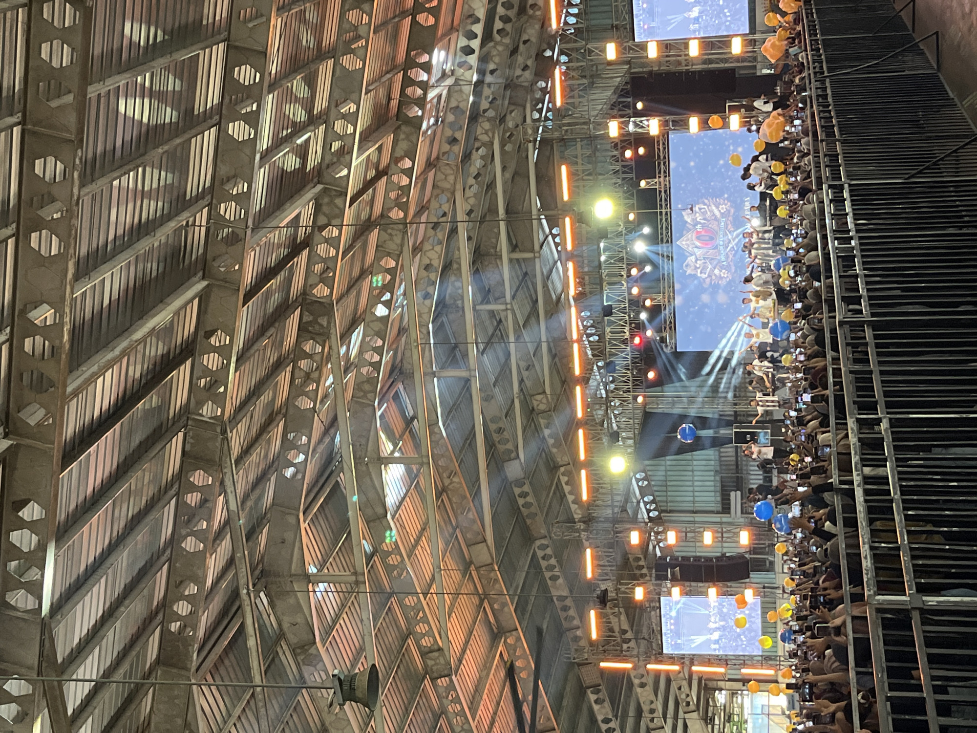 The image depicts a concert event taking place indoors, likely a large hall or stadium, with a visible corrugated metal ceiling and exposed structural beams. A stage is set up at the far end of the venue, featuring large LED screens displaying graphics and live feeds. A group of performers, dressed in white and black attire, are on stage, some holding instruments and interacting with the audience. Numerous orange and yellow lights illuminate the stage and the ceiling structure.
In the foreground, a large crowd of spectators is visible, many holding up their mobile phones to capture the event. The audience is separated from the stage by a metal barrier. Several blue and yellow balloons are scattered throughout the crowd and near the stage. The lighting suggests it is either evening or the venue has subdued natural light, with artificial stage lighting being the primary illumination. The overall atmosphere appears energetic and celebratory, typical of a live music performance. The location is specified as the city of Pelang, Indonesia.