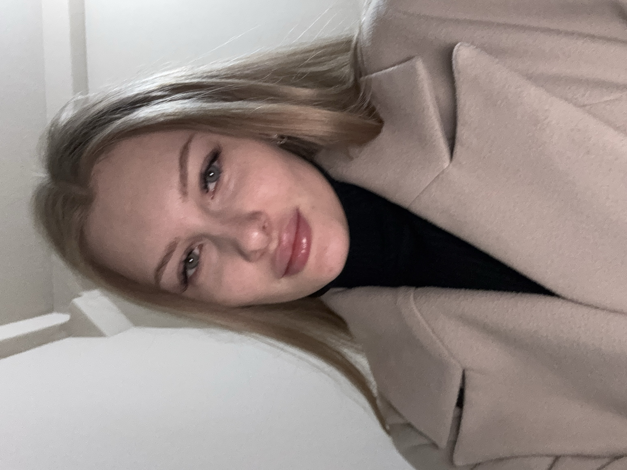 A light-skinned woman with blonde hair, wearing a black turtleneck and a tan-colored coat, poses for a selfie. The background appears to be a white or light-colored wall or interior surface. It is not possible to determine the location as Perm, Russia from the image alone.