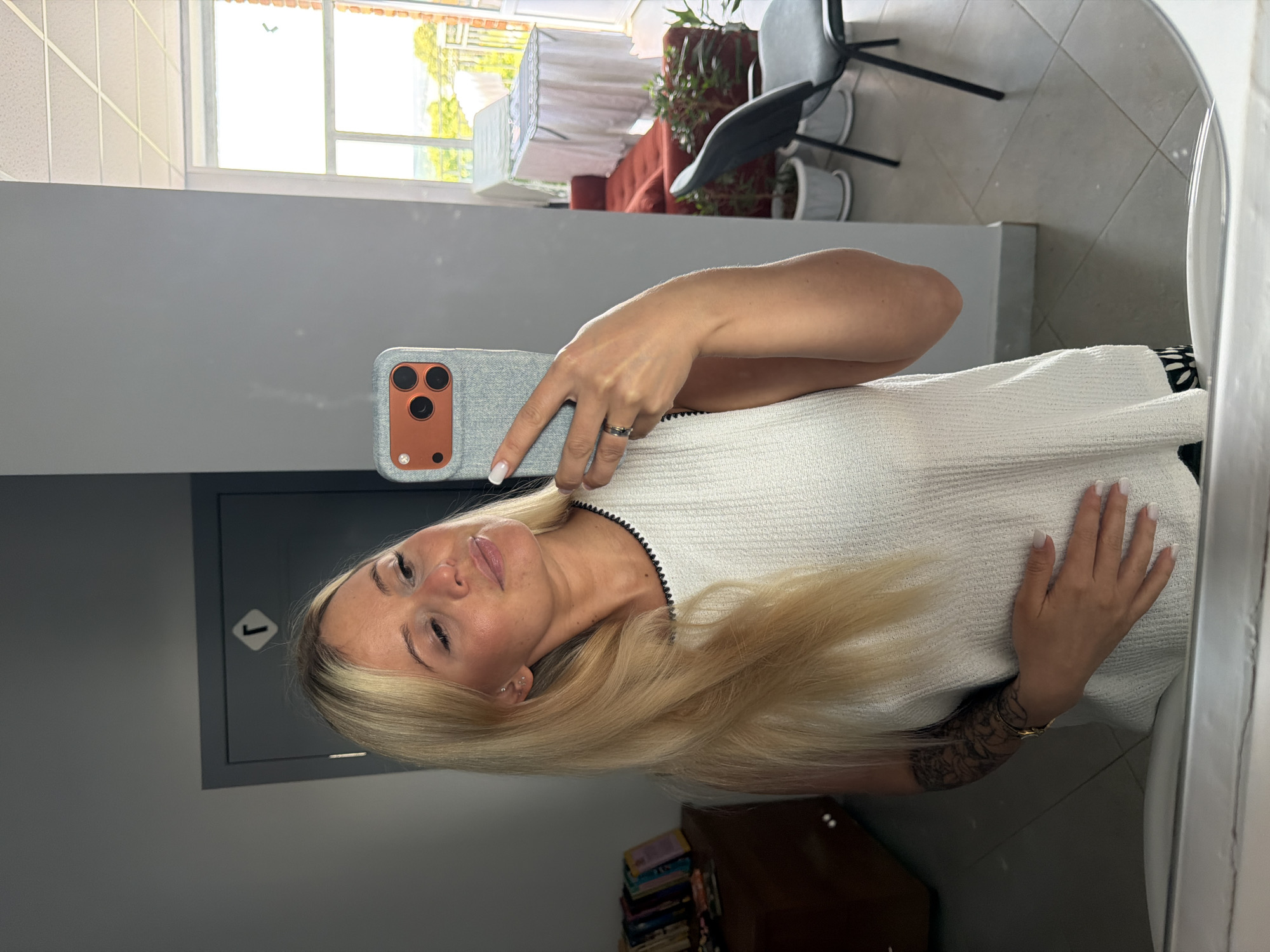 A woman with blonde hair and a white top is taking a selfie in a mirror. She is wearing a ring and has manicured nails. In the background, there is a window with sunlight streaming through, indicating it is daytime. The setting appears to be indoors, possibly a room with grey walls and tiled flooring. There are some plants and furniture visible in the reflection of the mirror. A phone case with a camera is visible in her hand. A sign with the number "1" in a diamond shape is on the wall behind her. The overall mood is casual and relaxed.