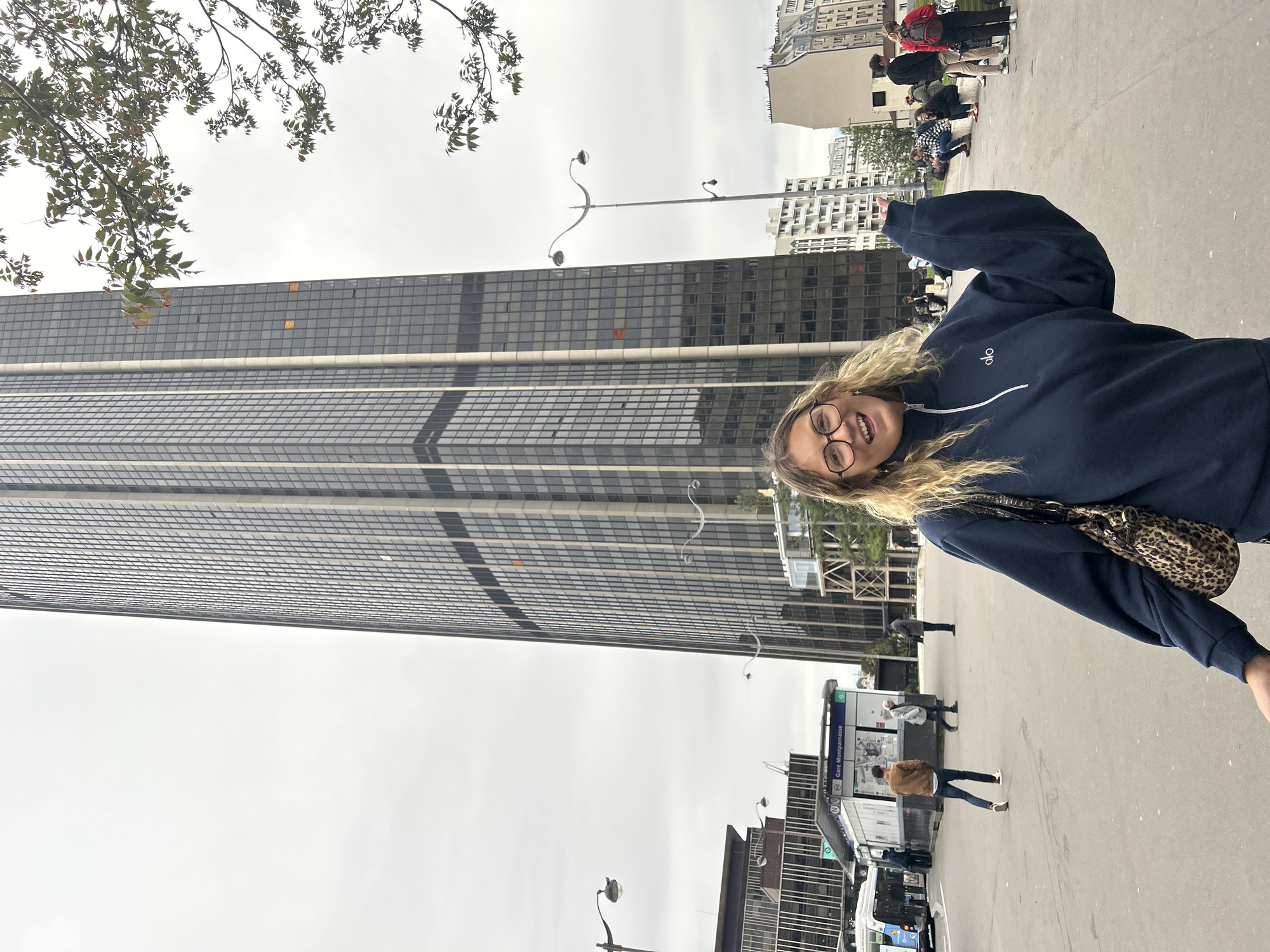 A woman stands on a city street with the Montparnasse Tower in Paris, France in the background. She wears a navy blue zip-up sweatshirt and glasses. The street is crowded with pedestrians. Public transportation is nearby. The sky is overcast.