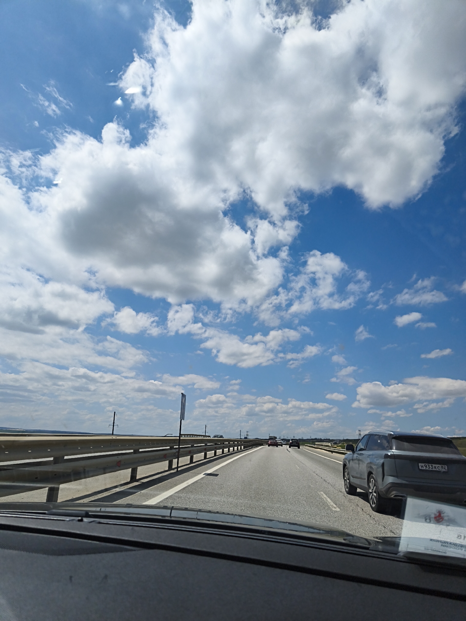 A highway stretches into the distance under a blue sky dotted with puffy white clouds. A gray car occupies the right lane, while a white line separates it from the adjacent lane. Several cars are visible further down the road. A metal guardrail lines the side of the highway. The view is from inside a car, with the dashboard visible in the foreground. The location is Unknown, Unknown.