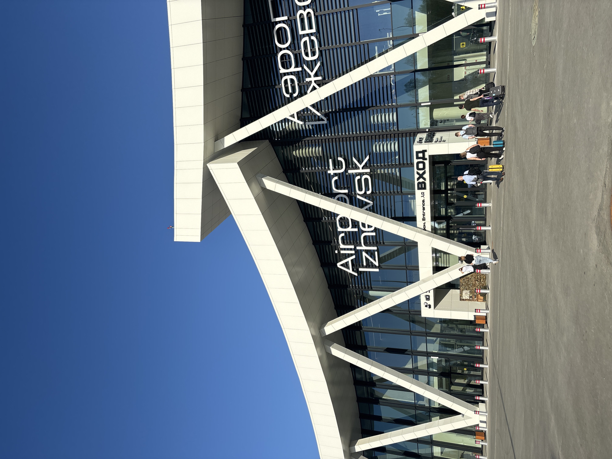 The image shows the modern facade of the Airport Izhevsk terminal in Новое Мартьяново, Russia. The building features extensive glass windows and a white structural support system. The airport name is prominently displayed in both English and Russian. A group of people with luggage is gathered near the entrance. The sky is clear and blue.