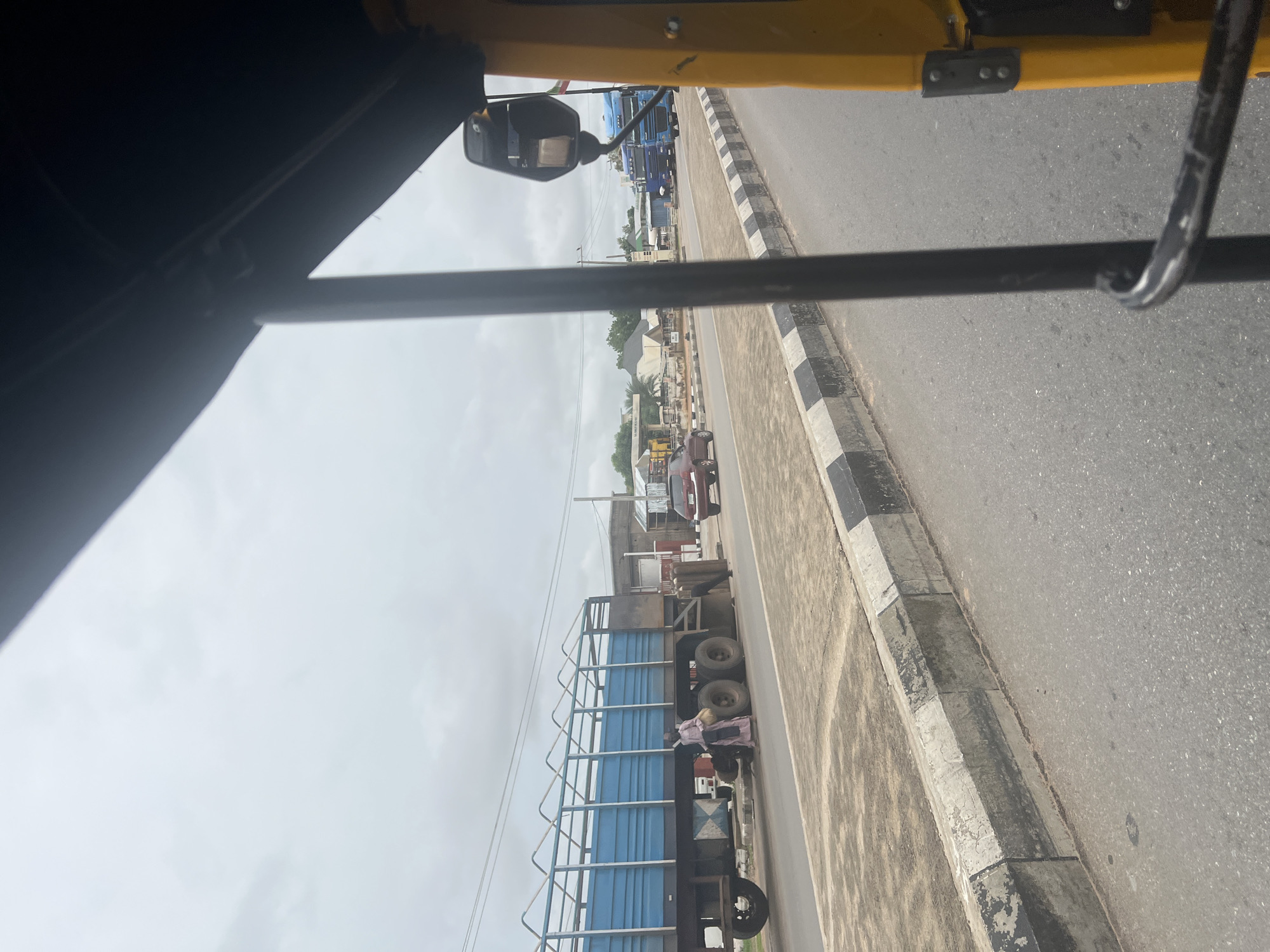 The image was taken from a yellow vehicle, possibly a tricycle or auto-rickshaw, looking towards a street scene in Jalingo, Nigeria. In the foreground, a black metal bar is visible, likely part of the vehicle's structure. The street is paved, with a raised concrete curb along the side, featuring black and white painted markings.
To the left of the street, a large blue and silver truck is parked. A person wearing a light-colored traditional garment is standing next to the truck, holding a light-colored bag or bundle. Another person is partially visible near the truck's rear. Beyond the truck, a red car is parked.
Further down the street, there are several buildings, including what appears to be a fuel station with a white canopy and pumps. Two blue semi-trucks are visible on the road in the distance. The sky is overcast with gray clouds. The overall environment suggests a commercial or roadside area within the city.