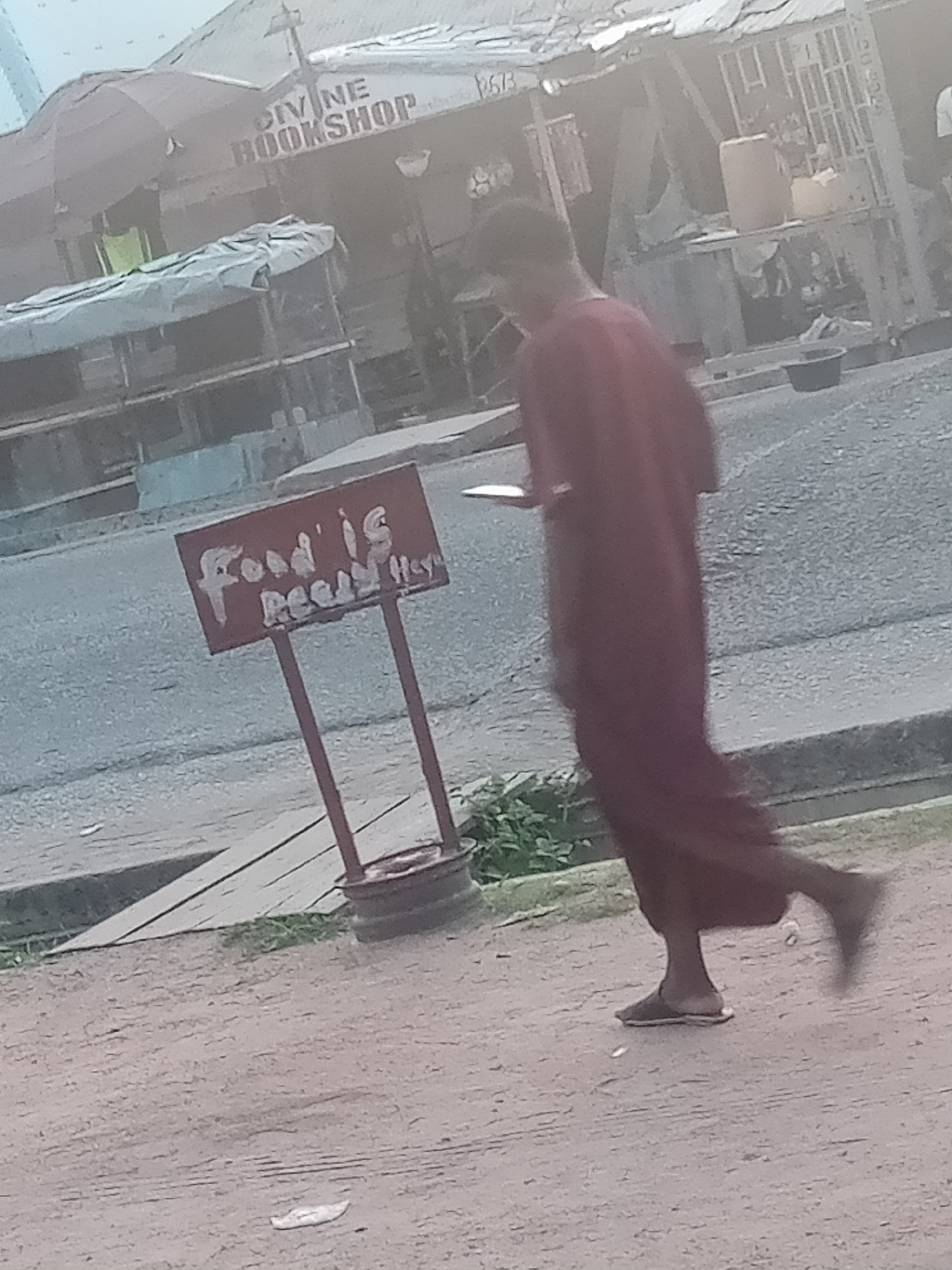 The image depicts a street scene in Jalingo, Nigeria, with a focus on a man in motion and a nearby sign. In the background, a structure with the words "DIVINE BOOKSHOP" is visible. The man, wearing a long, dark maroon garment and sandals, is walking or jogging to the right of the frame, with his left leg extended and his right leg bent. He appears to be holding a flat object in his right hand. In the mid-ground, a red sign with white lettering reads "FOOD IS READY". The sign is supported by two metal posts and rests on a circular metal base. The surrounding environment is an outdoor setting with a paved surface in the foreground and mid-ground, and a dirt or gravel path further back. Vegetation is visible to the left of the sign. The lighting suggests it is daytime, with some overcast conditions or shadows obscuring details.