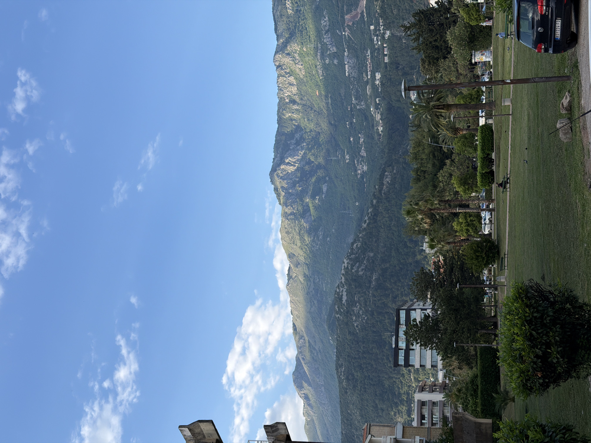 The image depicts a mountain backdrop with a grassy park area in the city of Budva, Montenegro. A portion of the sky is visible with scattered clouds. On the right of the frame is a section of a road with a parked black car. In the park, there are trees, bushes, light poles, and benches with a few people scattered around. Buildings are visible in the midground, nestled between the park and the mountain.
