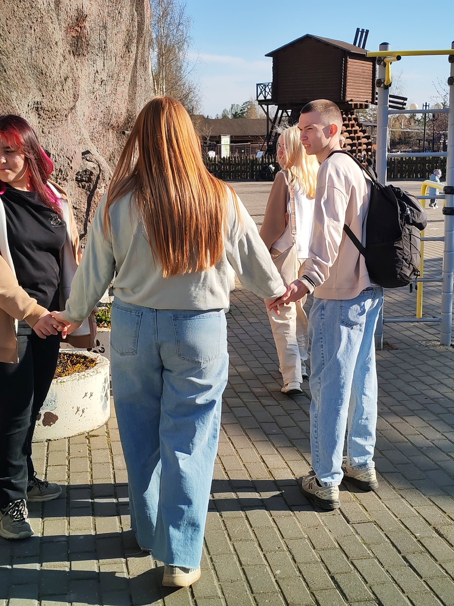Four young people are holding hands in a circle near a large artificial rock formation. The person closest to the camera has red and black hair and wears a black shirt, tan jacket, and black pants. The next person has long red hair, a light gray sweater, and wide-legged blue jeans. The third person has blonde hair and wears a white top, tan jacket, and light-colored pants. The fourth person has a shaved head, a light peach sweater, blue jeans, and a black backpack. The location appears to be a park in Izhevsk, Russia, based on the presence of a wooden playground structure in the background. The ground is paved with gray rectangular stones.