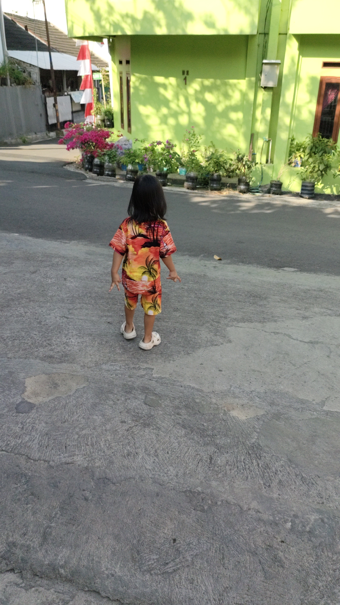 A young child, facing away from the camera, stands on a concrete sidewalk. The child is wearing a vibrantly colored tropical-themed outfit, consisting of a short-sleeved shirt and shorts, and white slip-on shoes. The child's back is to the viewer, and their hands are slightly extended to the sides.
Behind the child is a paved road, and further back, a bright lime green building. In front of the building, a row of potted plants, some with vibrant pink flowers, are arranged along the edge of the road. To the left of the building, a white wall with a red and white flag attached is visible. The scene appears to be a residential area within the city of Cimahi, Indonesia, possibly on a sunny day given the dappled light and shadows cast by trees on the building.