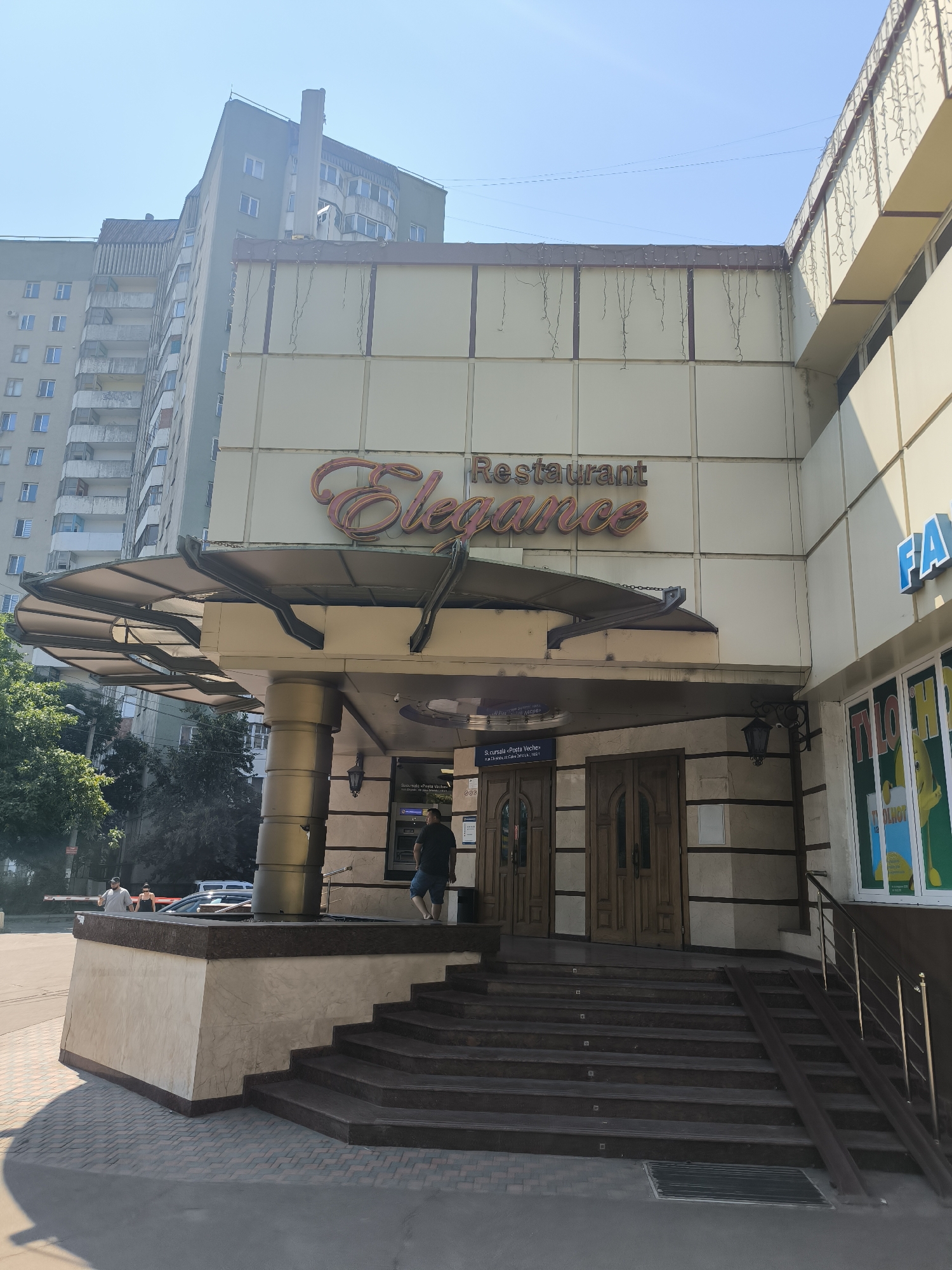 The image shows the entrance of a restaurant named "Elegance" in Chișinău, Moldova. The building has a modern facade with beige panels and decorative trim. Above the entrance, the restaurant's name is prominently displayed in a stylized font. A metal awning extends over the entrance, supported by columns. A set of stairs leads up to the double wooden doors of the restaurant. To the right of the entrance, there are windows displaying posters or advertisements. A ramp is also visible alongside the stairs, likely for accessibility. The street in front of the restaurant is paved, and there are trees and other buildings in the background, indicating an urban setting. Several people are visible near the entrance, including a man standing on the steps and others on the sidewalk. The sky is clear and blue, suggesting a sunny day.