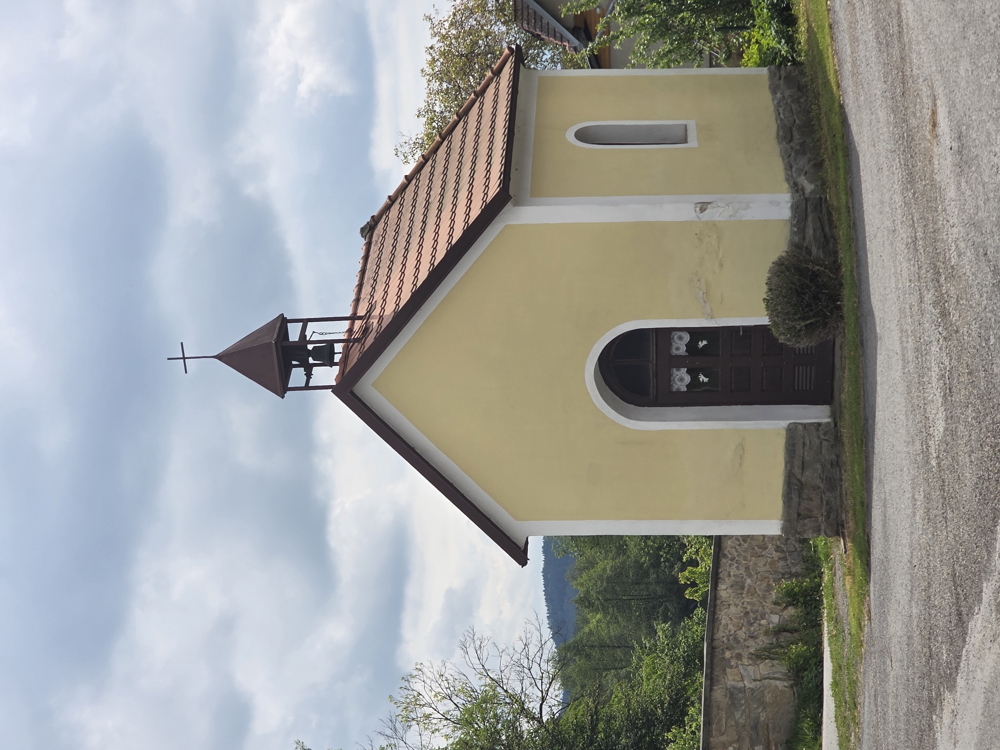 The image shows a small, yellow chapel with a red-tiled roof and a bell tower topped with a cross. The chapel has a white trim around the edges and windows, with a single, arched window above the door. The door is dark wood with decorative lace curtains visible in the arched opening. The chapel is located next to a paved road, with a low stone wall partially visible at the base of the building. Trees and greenery are visible in the background, suggesting a rural or semi-rural location. The sky is visible with some clouds.