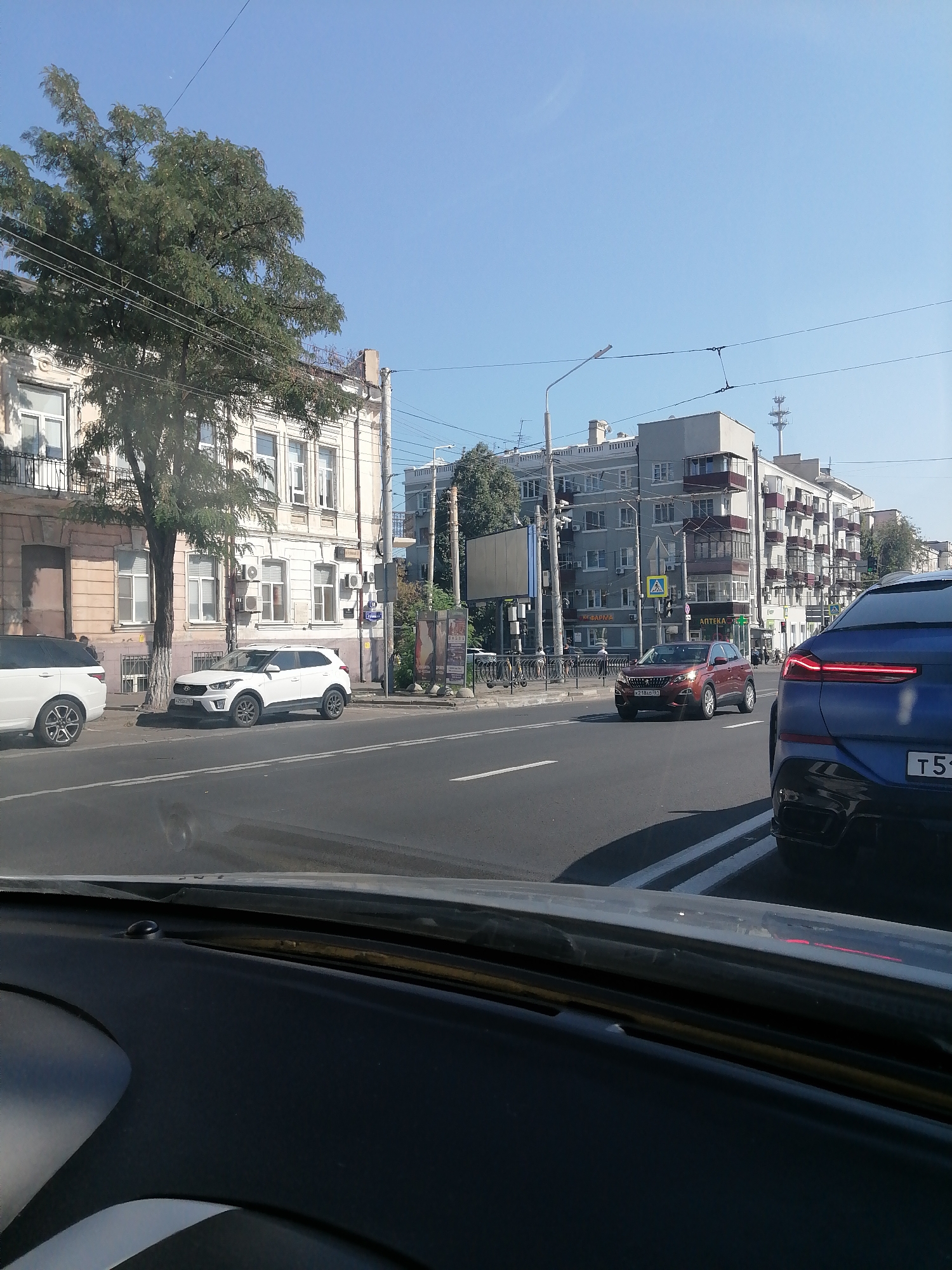 The image depicts a street scene in Rostov-on-Don, Russia, likely on a sunny day, given the clear blue sky. In the foreground, the interior of a car is visible, specifically the dashboard and part of the windshield. The street itself is paved with asphalt and marked with white lines indicating traffic lanes.
Several vehicles are present on the street. On the right side, a blue SUV is partially visible, its rear taillights illuminated. Further down the road, a red SUV is driving in the same direction as the blue SUV. On the left side of the street, parked along the curb, are a white Range Rover and a white Hyundai Creta.
The background features a mix of architecture. To the left, an older, light-colored building with multiple windows and balconies stands. To the right, modern apartment buildings with balconies and air conditioning units are visible. Streetlights and power lines are also present. A few trees with green foliage are scattered throughout the scene, adding to the urban environment. The overall impression is a typical city street with traffic and residential buildings.