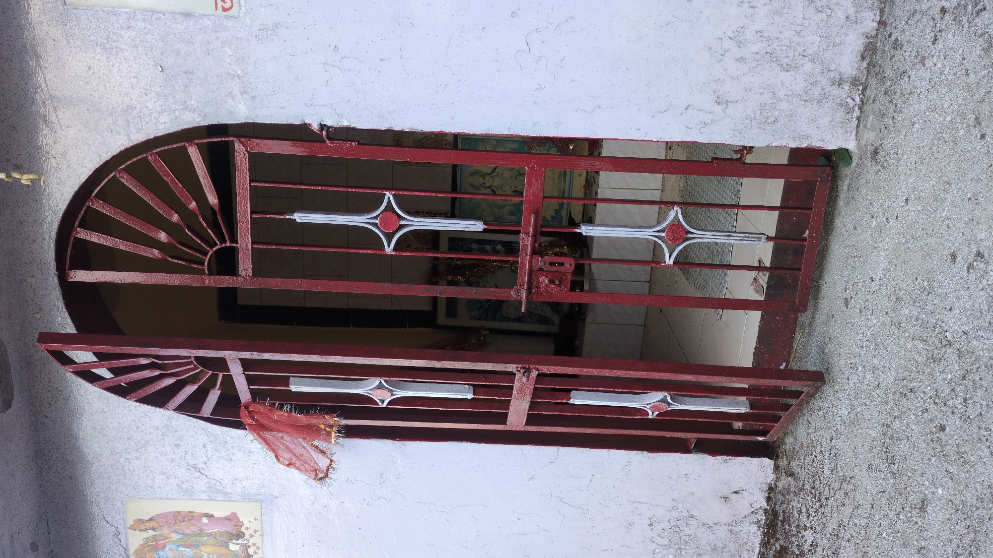 A barred window is set in a white wall. The bars are painted maroon, and have decorative white and red accents. A small scrap of red cloth is tied to the lower left corner of the bars. Through the bars, one can see the interior of a room with tiled walls. Religious images are visible inside. The wall surrounding the window is cracked and stained. The location is Haini, India.