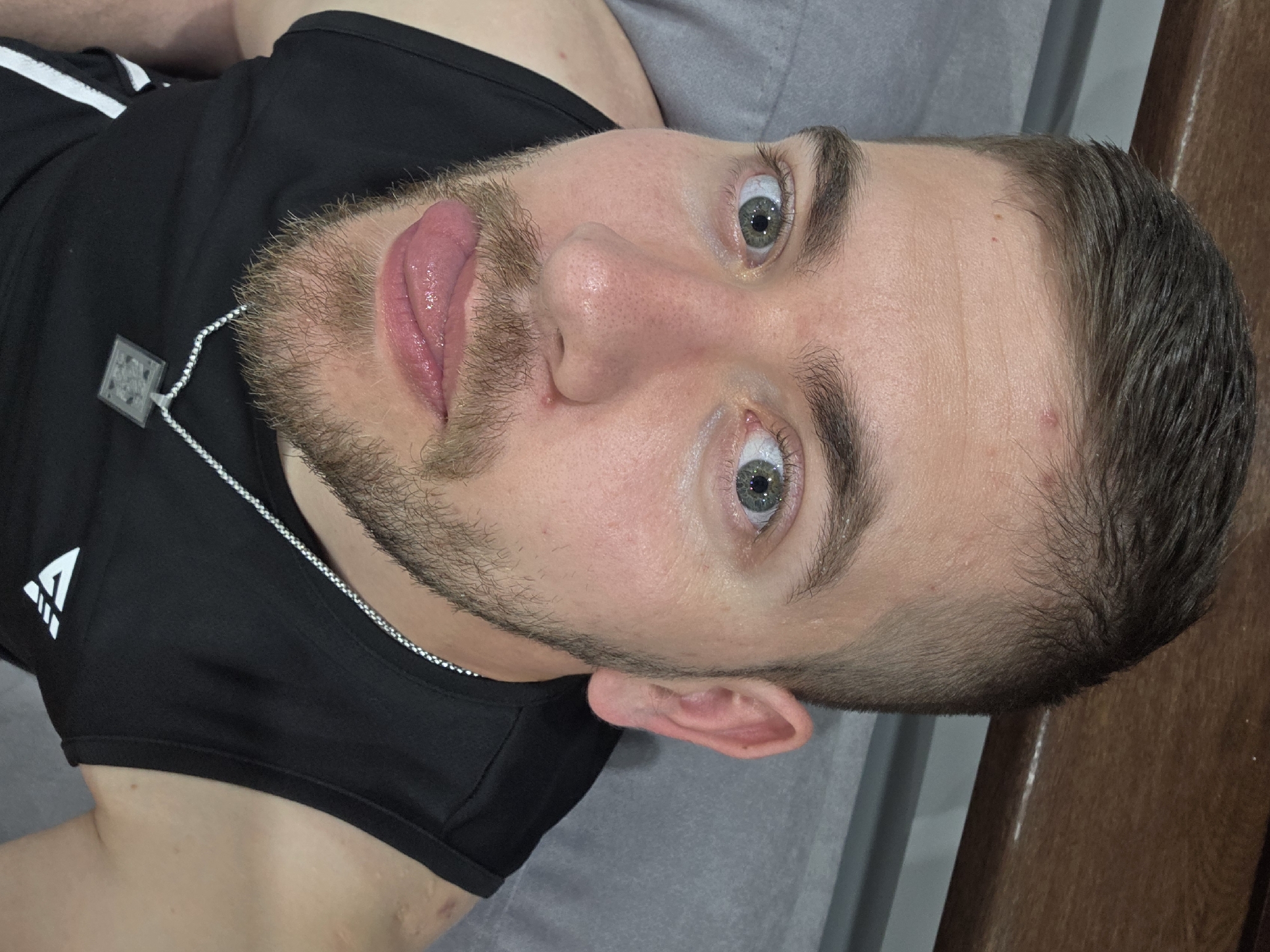 A young man is depicted in a close-up, portrait-style image, lying down and looking towards the camera. He has short brown hair, a light beard and stubble, and striking light-colored eyes (appearing greenish-blue). His tongue is extended to the side, creating a playful or quirky expression. He is wearing a black athletic tank top with a white logo on the left chest, which consists of an abstract triangular shape with three horizontal lines. A thin silver chain with a square, possibly metallic, pendant is visible around his neck.
Notable details include a few small blemishes or moles on his forehead and cheek, and what appears to be a minor scar on his left shoulder/neck area. His pupils appear dilated. The man is positioned on a soft, gray surface, likely a bed or couch. In the background, to the right, a darker brown surface, possibly wooden furniture or a wall, is visible. The lighting is bright and even, highlighting his features. No specific time of day or weather can be determined from the indoor setting, but the image is well-lit. The overall impression is an informal, personal photo. This image is set in Yagodovo, Bulgaria.