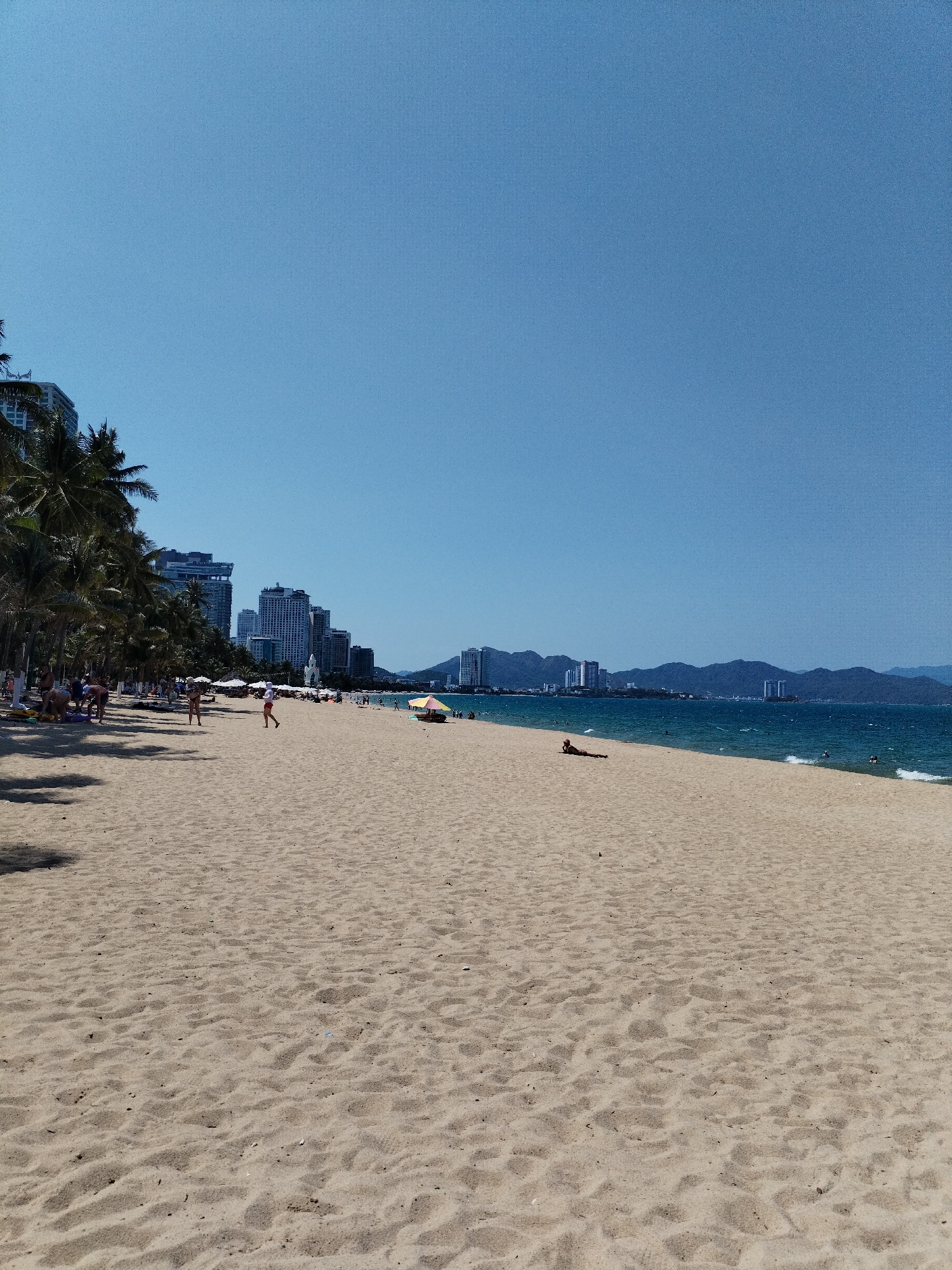 The image shows a wide, sandy beach on a sunny day in Nha Trang Ward, Vietnam. The foreground is dominated by light-colored sand, marked by footprints and patterns. Along the left side of the frame, a line of palm trees provides shade for beachgoers. Behind the trees, modern high-rise buildings are visible, indicative of a coastal city. In the distance, a mountain range forms the horizon. The sea is a mix of blue and green. A few people are scattered across the beach: some are sunbathing, others are walking, and a few are in the water. A small, colorful beach umbrella is set up in the middle distance. The sky is a clear, bright blue.