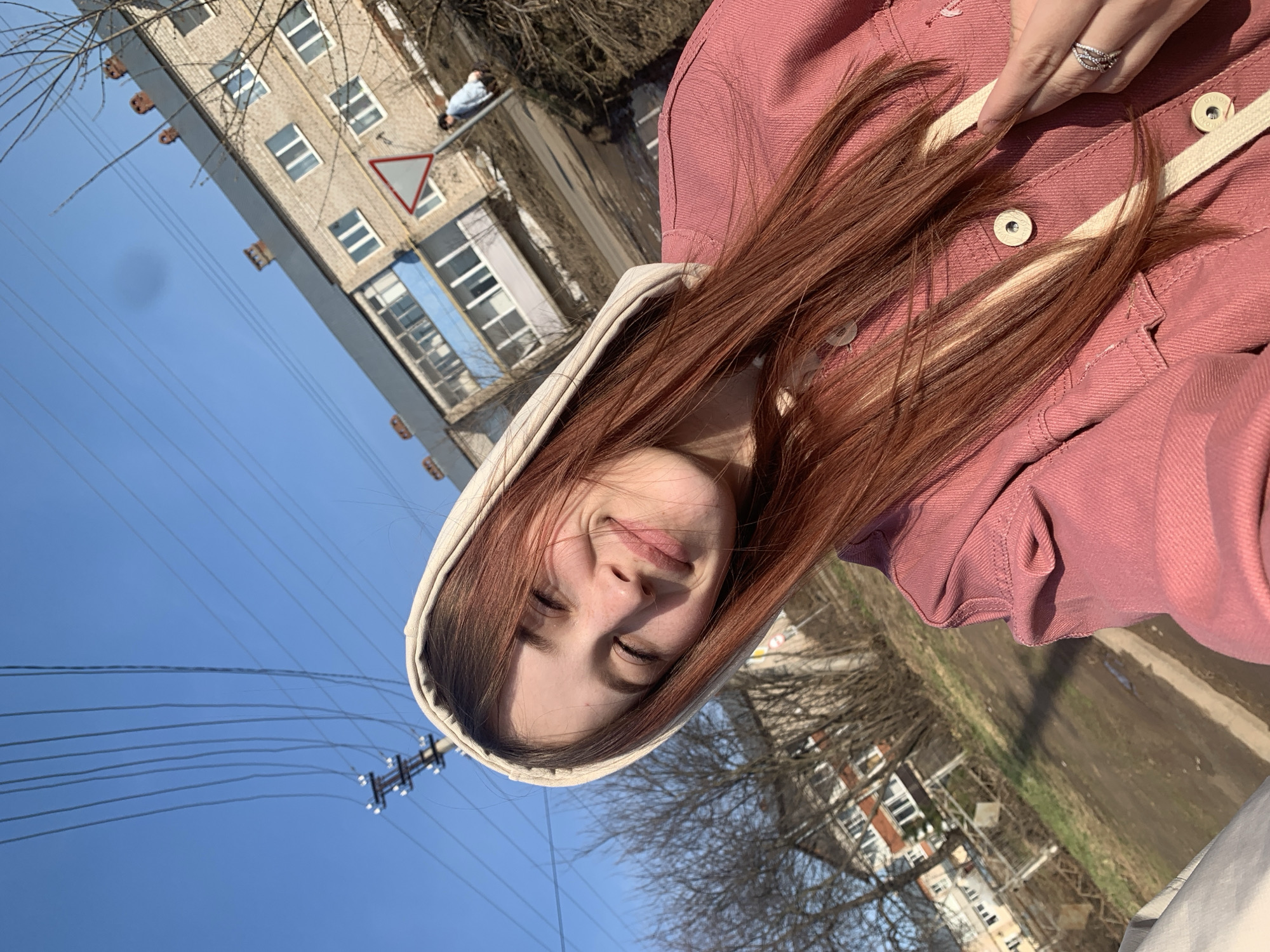 A young woman is photographed in a selfie, outside in Фоки, Russia. The perspective is low and upward, the camera tilted. She wears a pink hooded jacket with a cream-colored hood lining and drawstrings. She has red-brown hair and is smiling with her eyes closed. She wears a ring on her left hand. The background shows a clear blue sky, telephone poles with wires, and a multistory apartment building. There is a street sign next to the building. The ground is visible with some bare trees and other buildings. The photo is taken during the day in possibly the spring or fall.