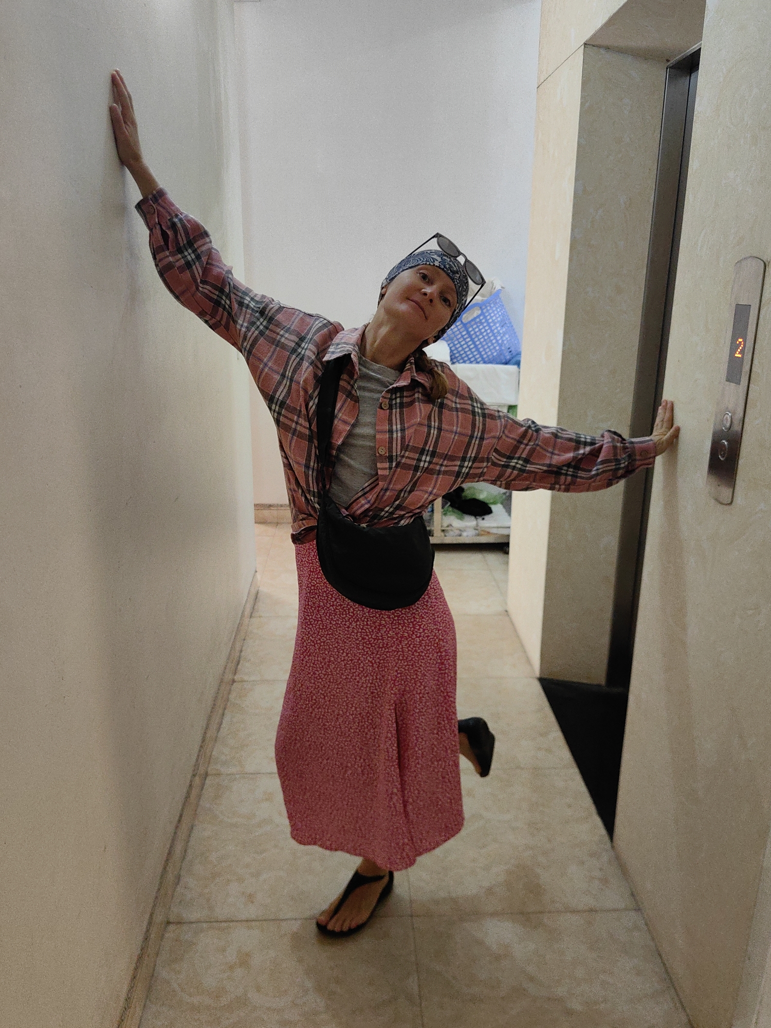 A woman stands in a narrow hallway of a building in Bai Chay Ward, Vietnam, posing between a wall and an elevator. The elevator indicates floor "2". The woman is wearing a red patterned skirt, a gray t-shirt, a red plaid shirt tied at the waist, a black fanny pack, flip-flops, a headscarf, and sunglasses on her head. She has one hand on the wall and the other on the elevator panel. The hallway has tiled flooring. A glimpse into a room with a blue basket can be seen at the end of the hallway.