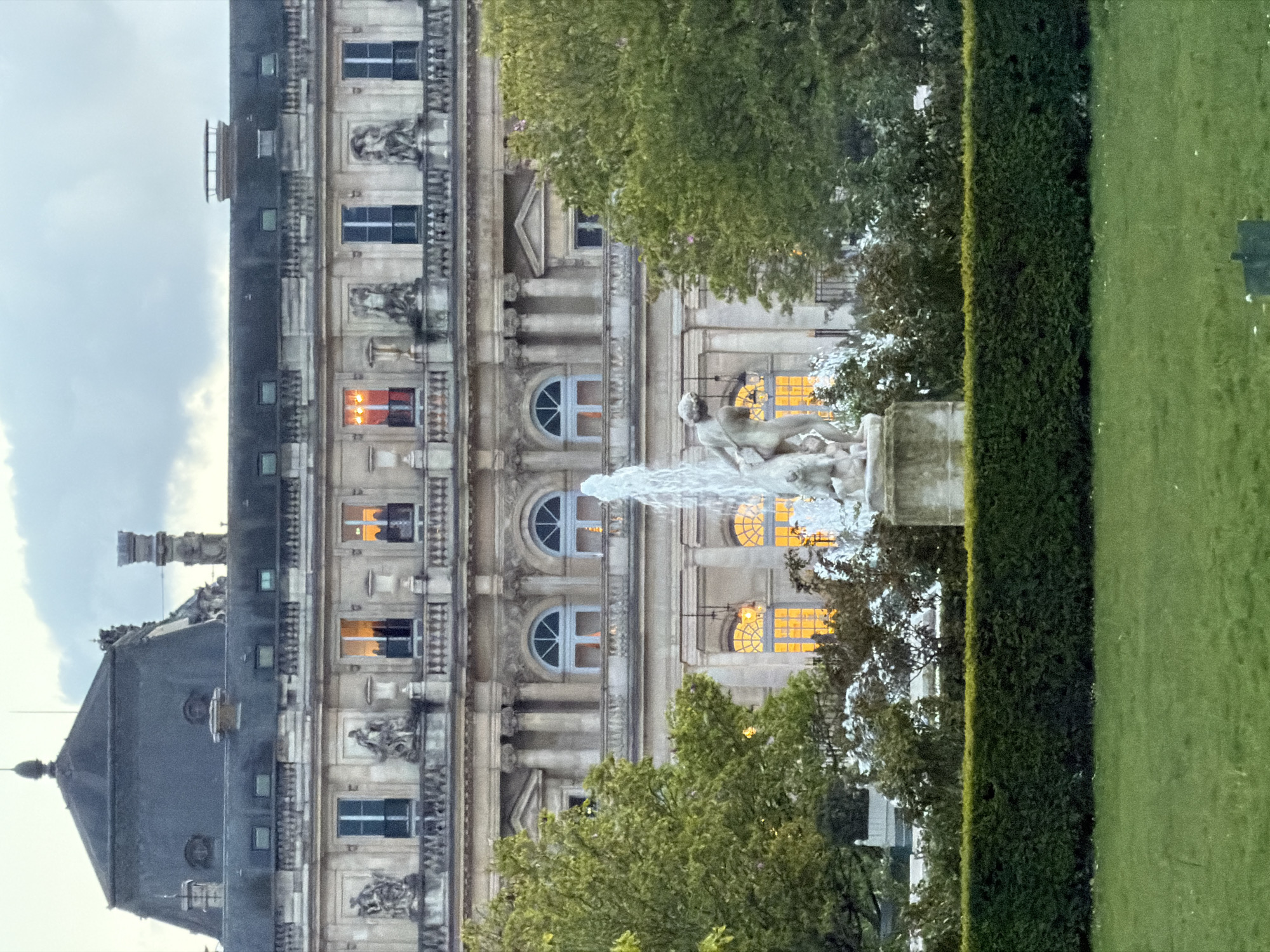 The image presents a frontal view of the Luxembourg Palace, located within the Luxembourg Garden in Paris, France, under an overcast sky. In the immediate foreground, a segment of green grass is visible, followed by a neatly trimmed, dark green hedge. Behind the hedge, a stone fountain is centrally positioned, actively spraying water upwards from a central sculpture. This sculpture depicts a nude male figure standing next to a smaller, indeterminate animal or cherub-like form.
The midground features various green trees and shrubs, partially obscuring the lower section of the palace. The background is occupied by the multi-story Luxembourg Palace, constructed of light-colored stone. Its classical facade includes multiple rectangular and arched windows across several levels. Several windows on the visible floors emit a warm, golden light from within. The architecture displays classical columns, balustrades, and sculptures integrated into the upper facade. The palace is topped by a dark grey mansard roof, featuring several dormer windows and a pointed finial at its apex. No human figures are present in the scene.