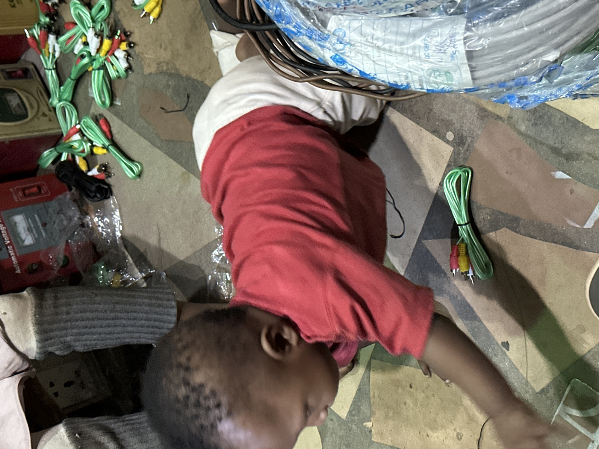 In Barden Yana, Nigeria, a young child, likely a toddler, is crawling on a concrete floor. The child is wearing a red long-sleeved shirt and off-white pants. In front of the child, on the floor, are several bundles of green AV cables with red, yellow, and white connectors. Also visible are various electronic components and packaging, suggesting a commercial or storage area. The background shows what appears to be an electrical outlet and possibly electrical equipment. The lighting is dim, contributing to a somewhat cluttered and dimly lit environment.