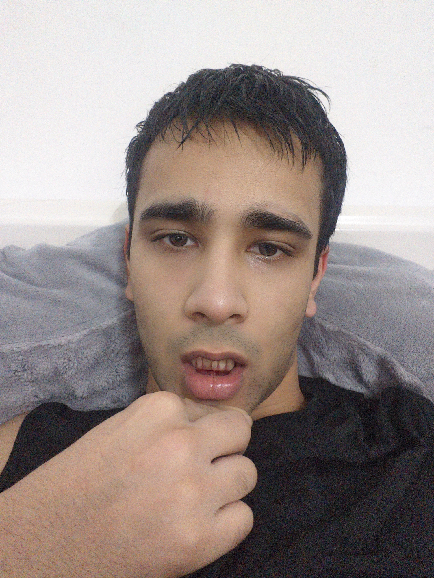 A medium close-up shot of a young man with dark hair and brown eyes. He is fair-skinned, and his hair appears slightly wet. He is wearing a black T-shirt and is resting against a grey pillow or soft surface. His right hand is raised to his chin, with his fingers lightly touching his jawline. The background is a plain white wall.
There is no information in the image to determine a location or city.