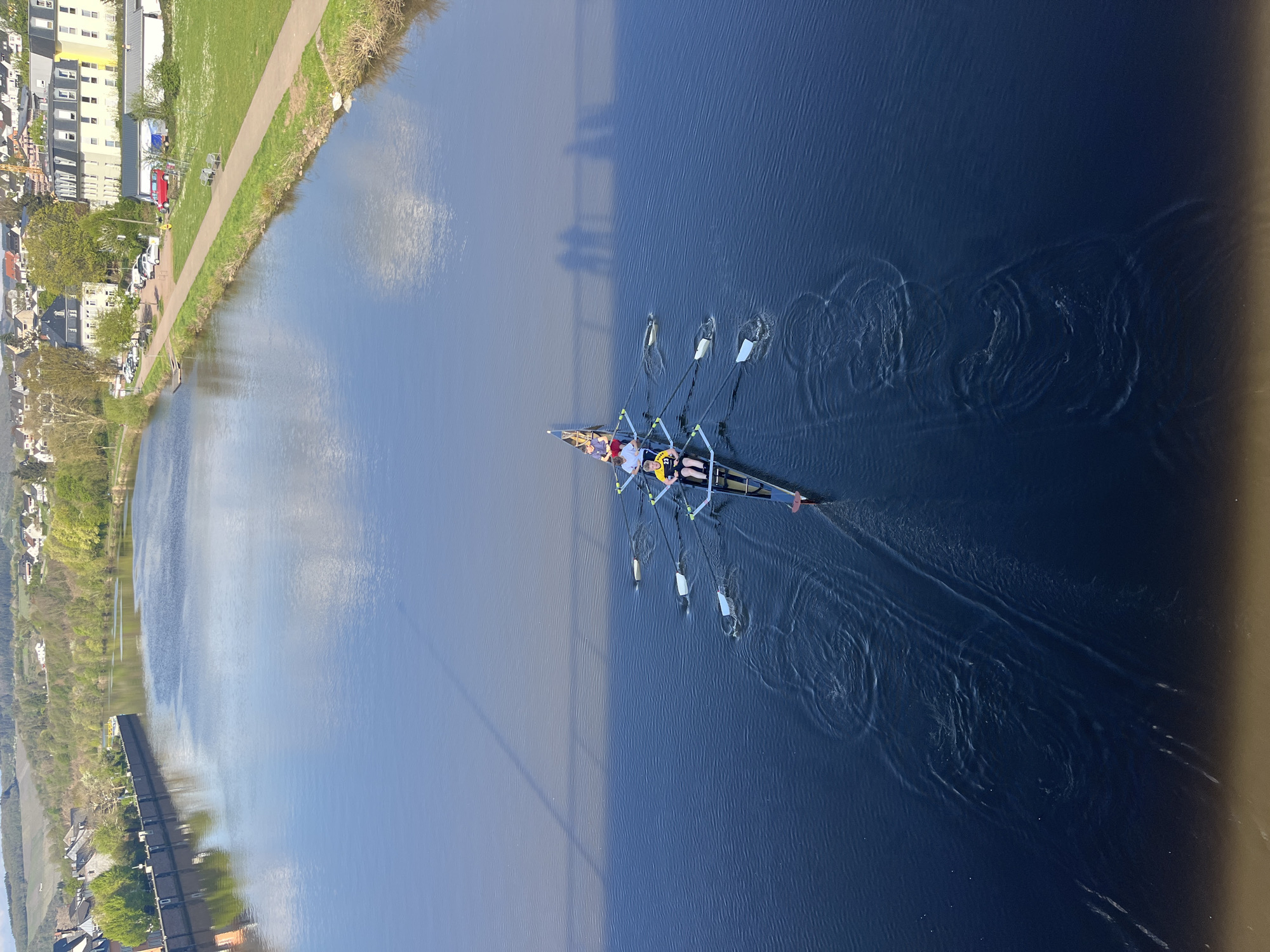 An aerial, eye-level image shows a rowing crew team in a river in Saarburg, Germany. The team of eight rowers, plus a coxswain, is rowing an eight-person shell. The rowers are wearing various athletic apparel, including yellow, white, red and blue colored shirts. The oars are dipping into the water, creating circular ripples. The river reflects the sky with some cloud cover. The riverbanks are lined with buildings, trees, and a walking path. The image is taken from above the river, potentially from a bridge. The shadow of the bridge is visible.