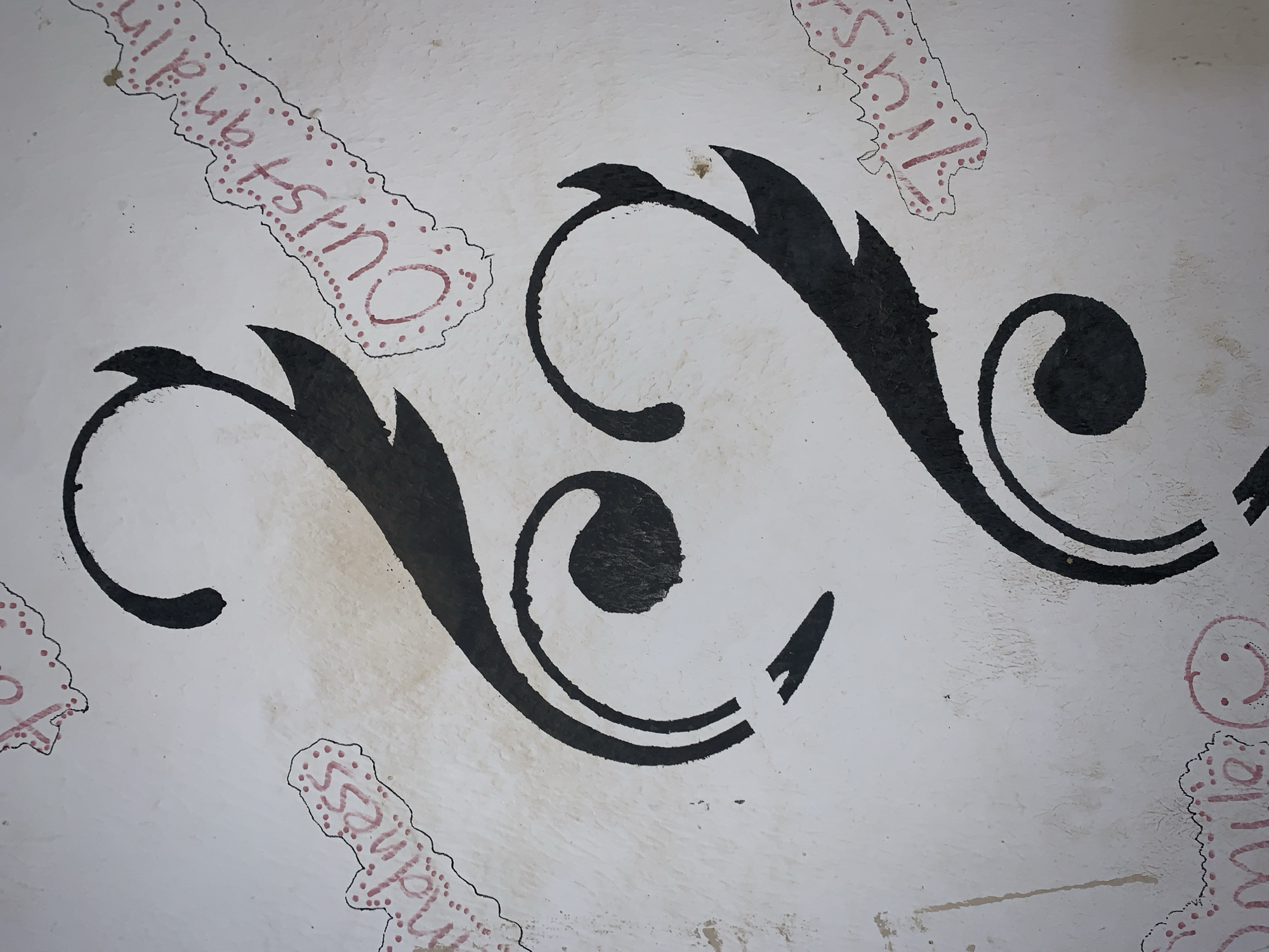 The image shows a close-up of a surface, possibly a wall or canvas, painted white with decorative elements in black and pink. The dominant elements are two large, stylized floral or vine-like designs in black. Superimposed over the white background are irregular, elongated shapes outlined in black with pink lettering and small dots within, spelling out words like "Outstanding," "Kindness," and "Smile," along with a smiley face symbol. The location is Rafin Par, Nigeria. No people are visible.