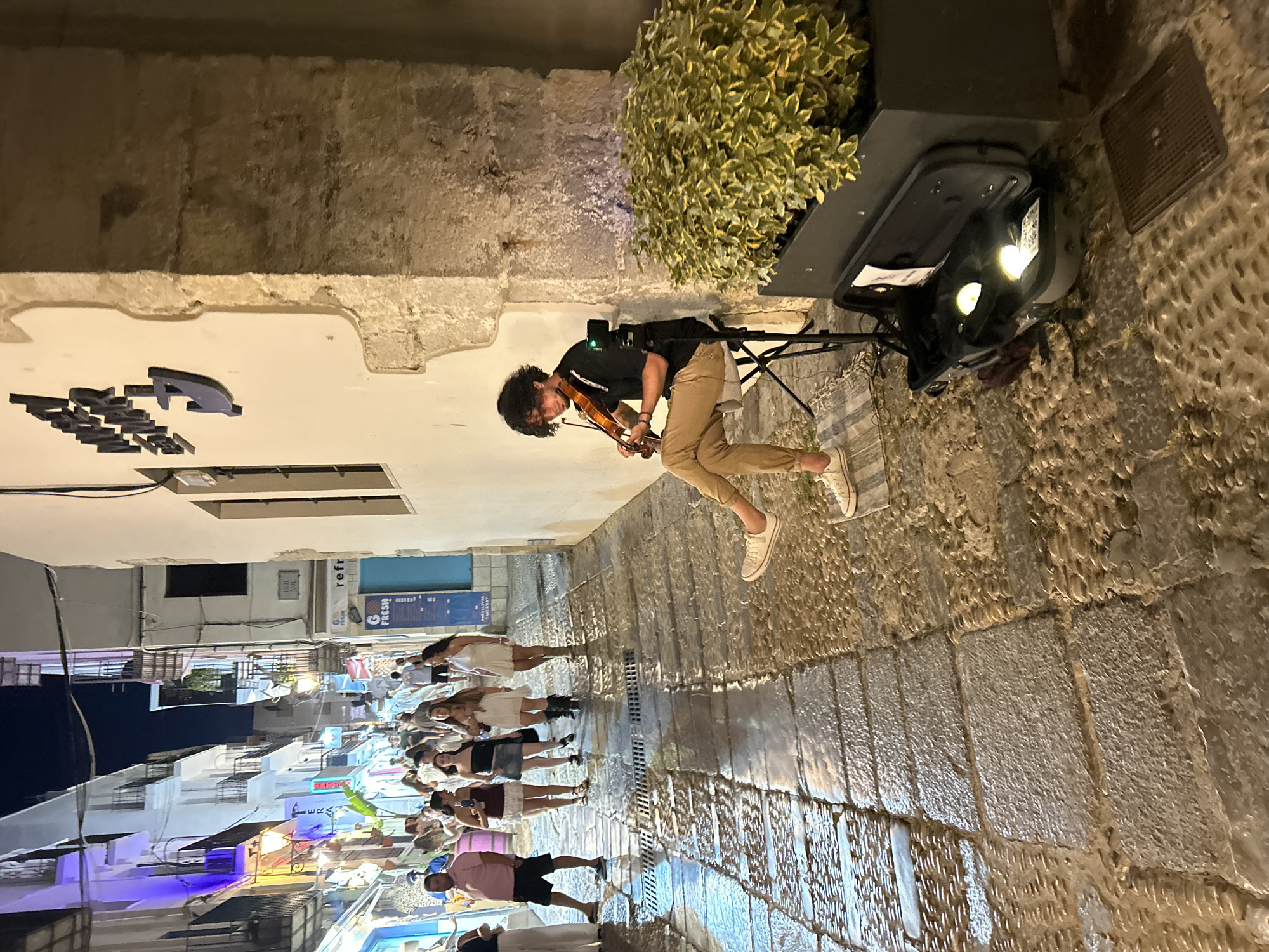 An evening street scene in the historic district of Peníscola, Spain, captures a young male street musician playing the violin. The narrow, cobblestone street is wet, reflecting the ambient light from the surrounding buildings and street lamps. Buildings on either side of the street are illuminated with colorful lights, showcasing their architecture and balconies.
In the foreground, the violinist is seated on a folding chair, dressed in a black shirt and tan pants, his violin resting on his shoulder. A black violin case is open at his feet, containing two circular lights. A small video camera is mounted on a stand behind him.
Further up the street, a group of people are walking, some appearing to be tourists. The overall atmosphere suggests a lively evening in a popular tourist destination. The architecture and the presence of the sea, hinted at by the name "MAR" on the wall, suggest the location is near the coast. The style of buildings, the cobblestone streets, and the general ambiance are characteristic of the old town of Peníscola, particularly the streets leading towards the castle.